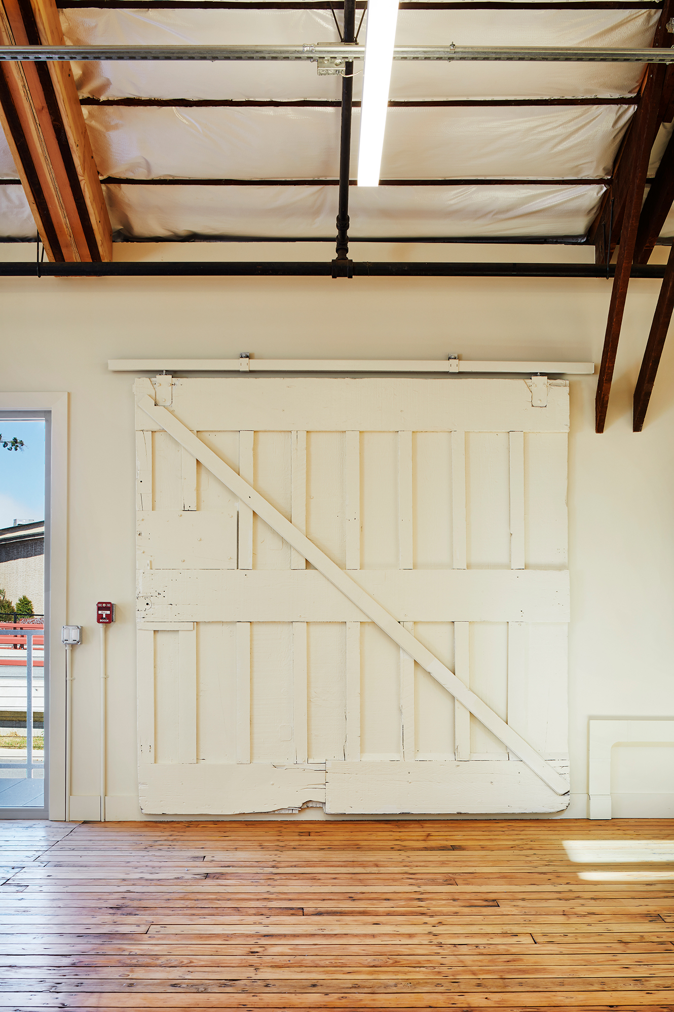 Large white sliding barn door mounted on exposed hardware inside a restored timber building with wood floors, visible roof insulation, and a suspended linear light.