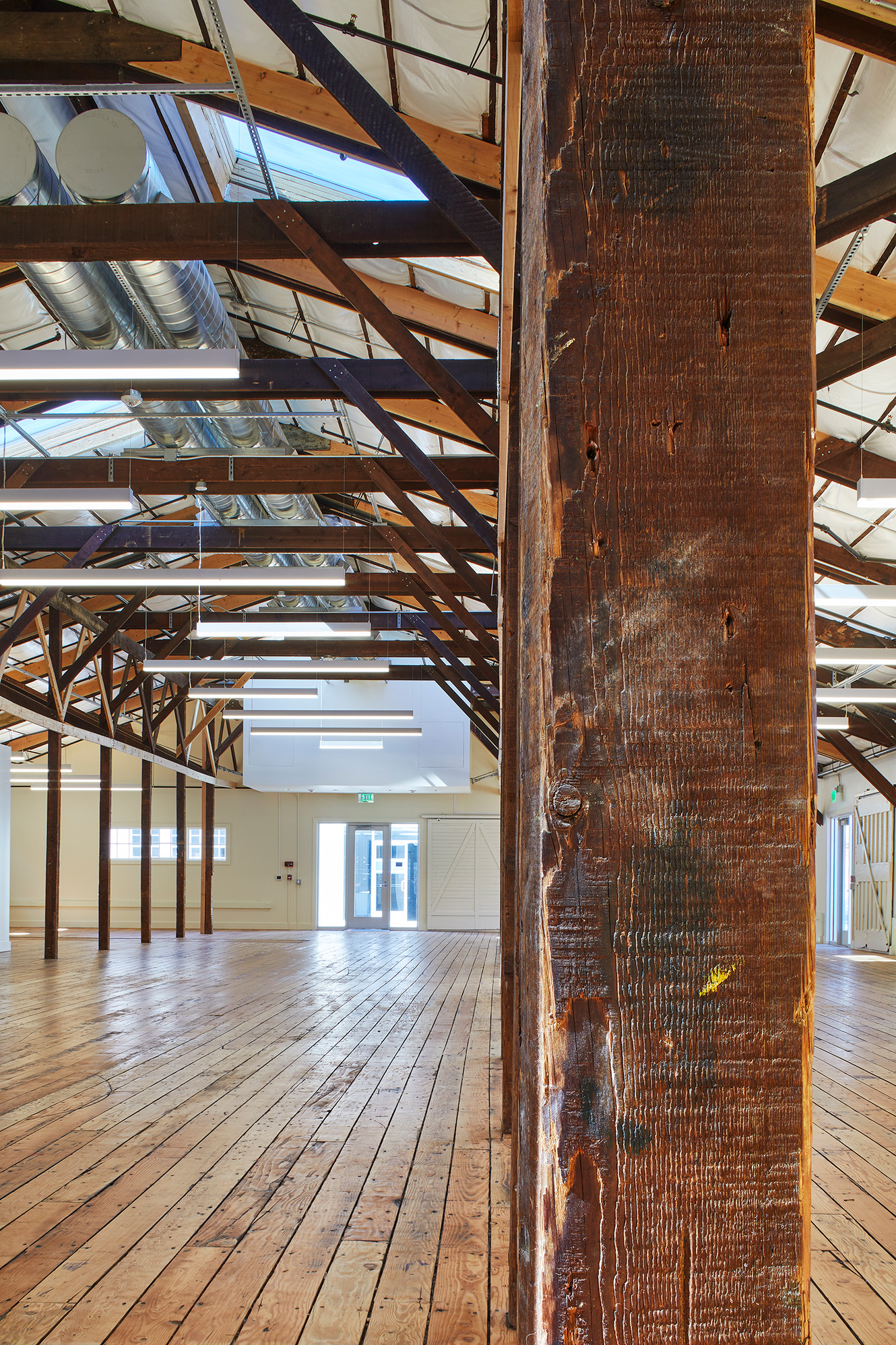 Open historic interior with restored wood floors, exposed timber trusses, suspended linear lights, and visible ductwork, framed by a close-up of a weathered wood column.