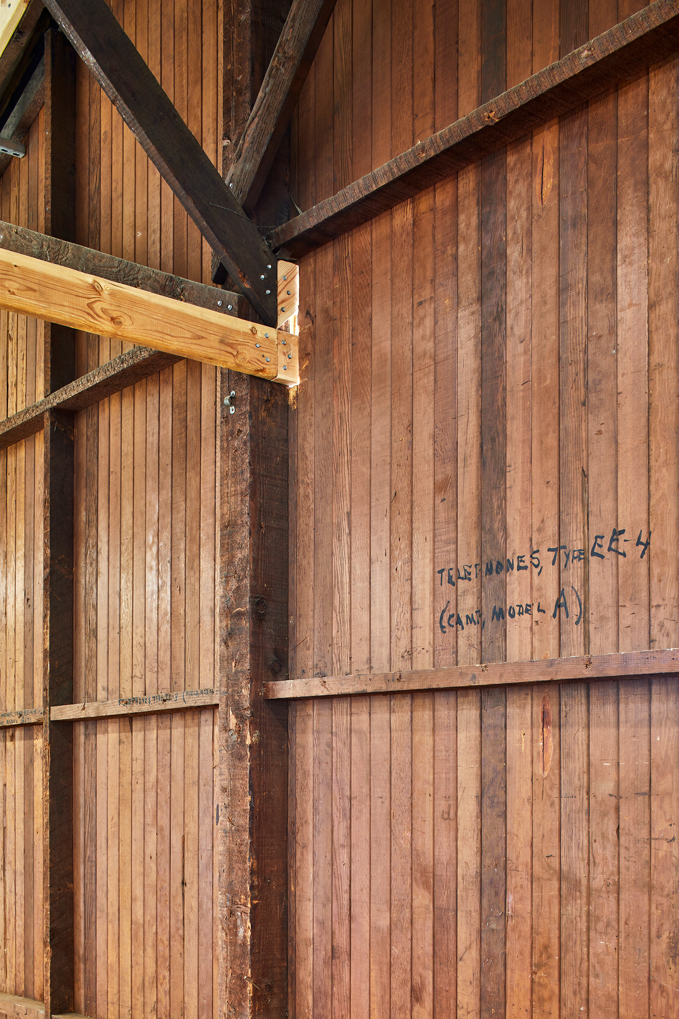 Close-up of historic wood wall framing and diagonal braces, with weathered boards, newer reinforcement members, and handwritten construction markings.