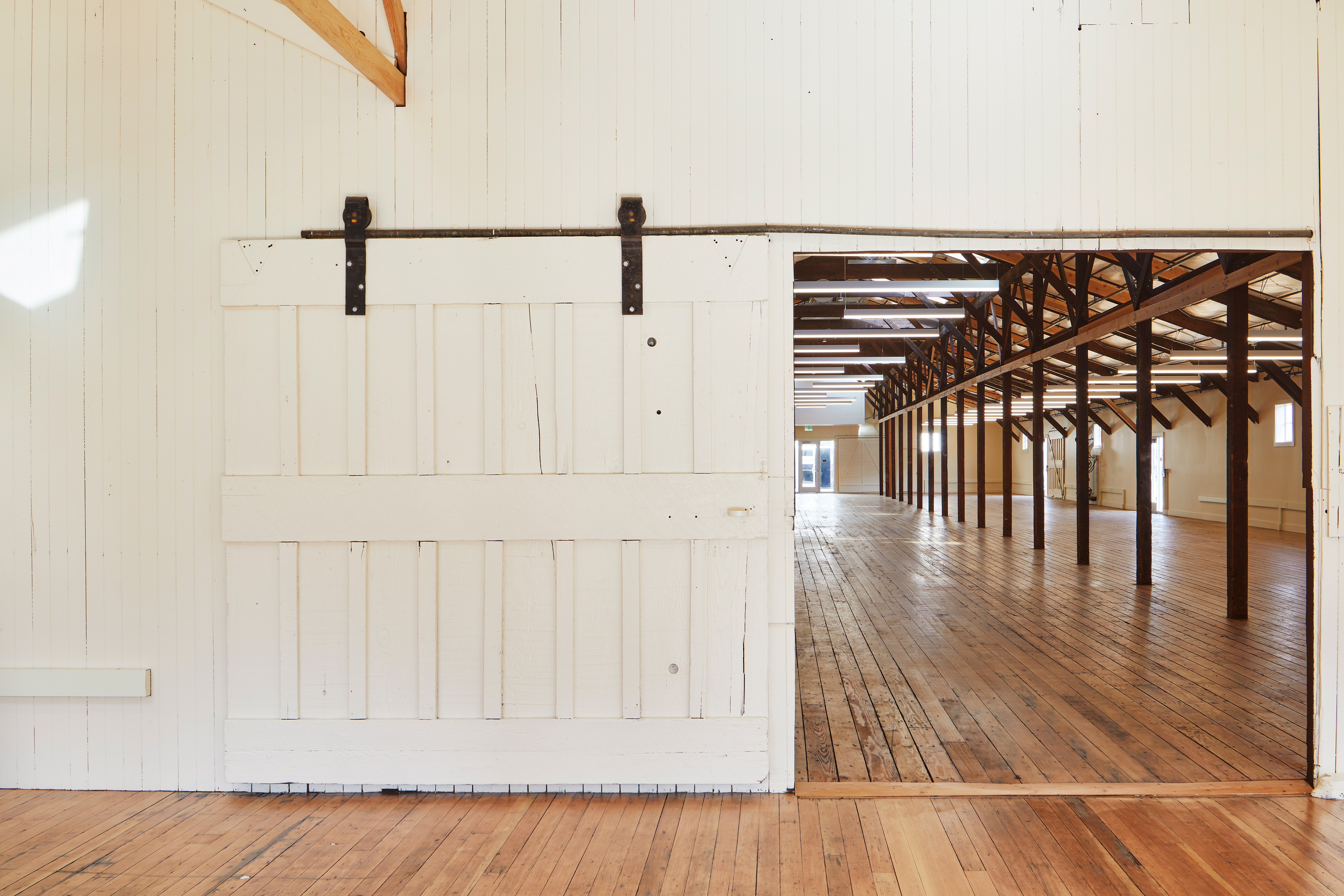 Large white sliding barn door opening into a long timber-framed interior with restored wood floors and suspended linear lights.
