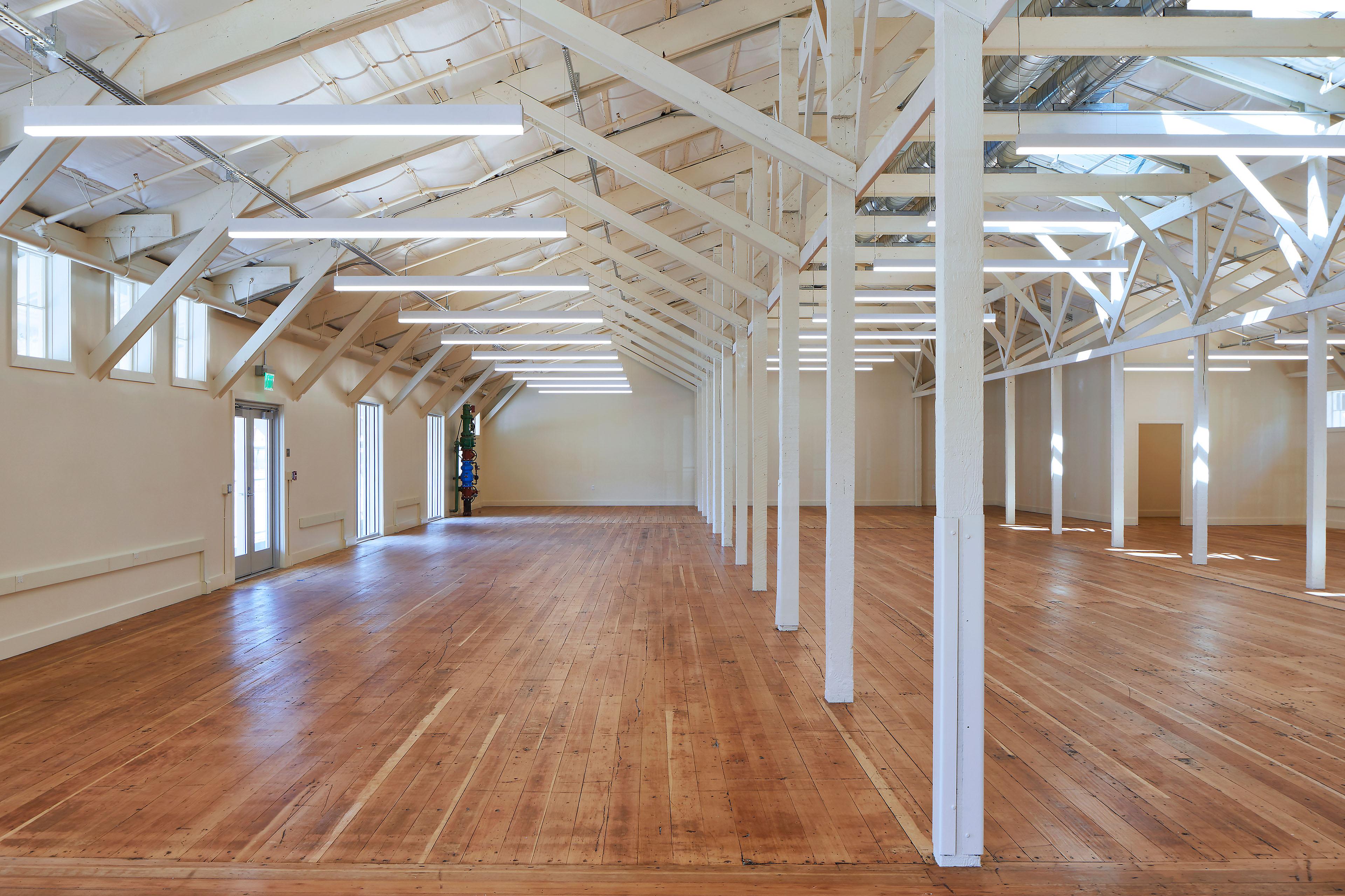 Expansive restored interior with wood floors, white-painted timber columns and trusses, suspended linear lights, and tall windows along the perimeter.