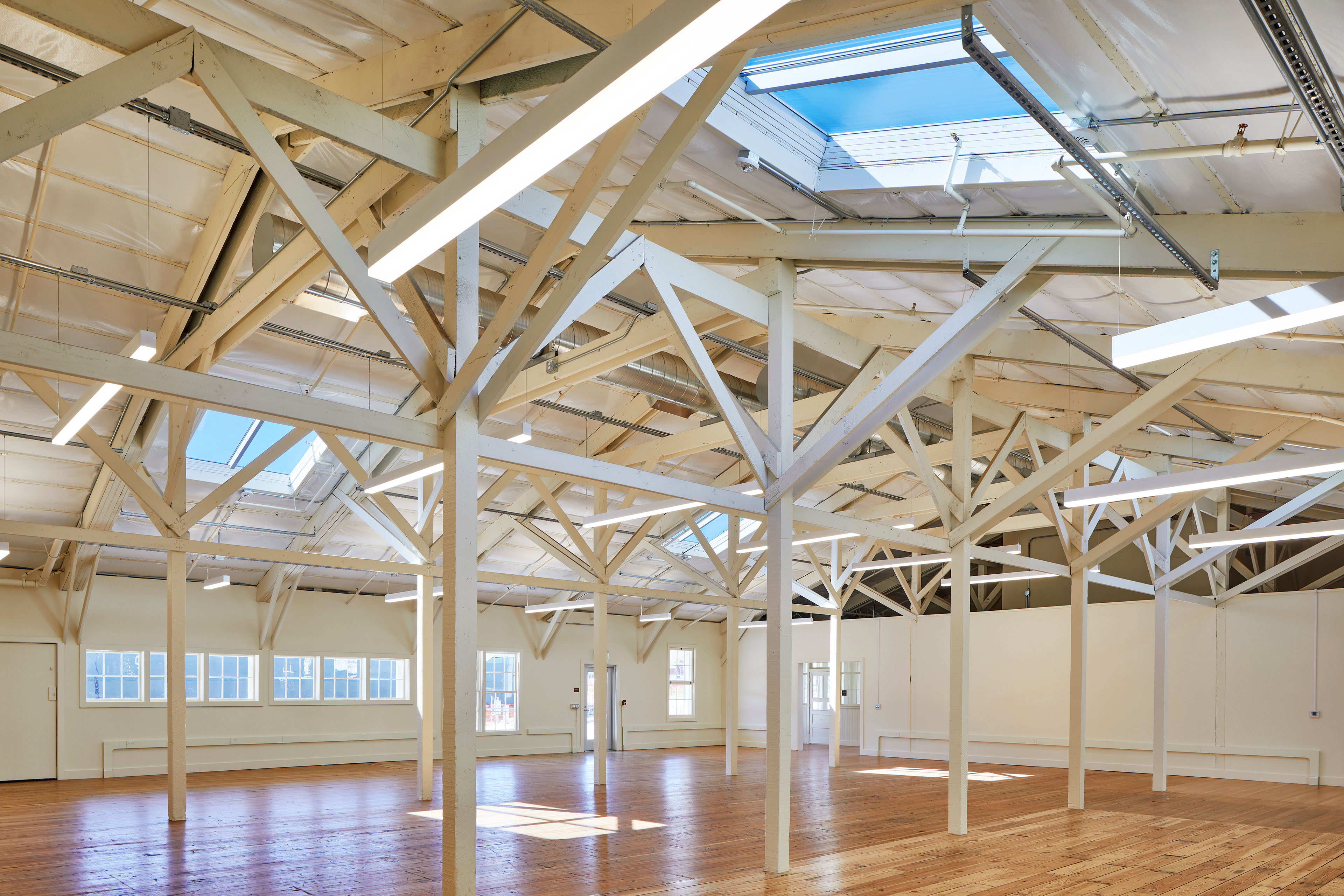 Open timber-framed interior with restored wood floors, white-painted structural framing, suspended linear lights, and skylights bringing daylight into the space.