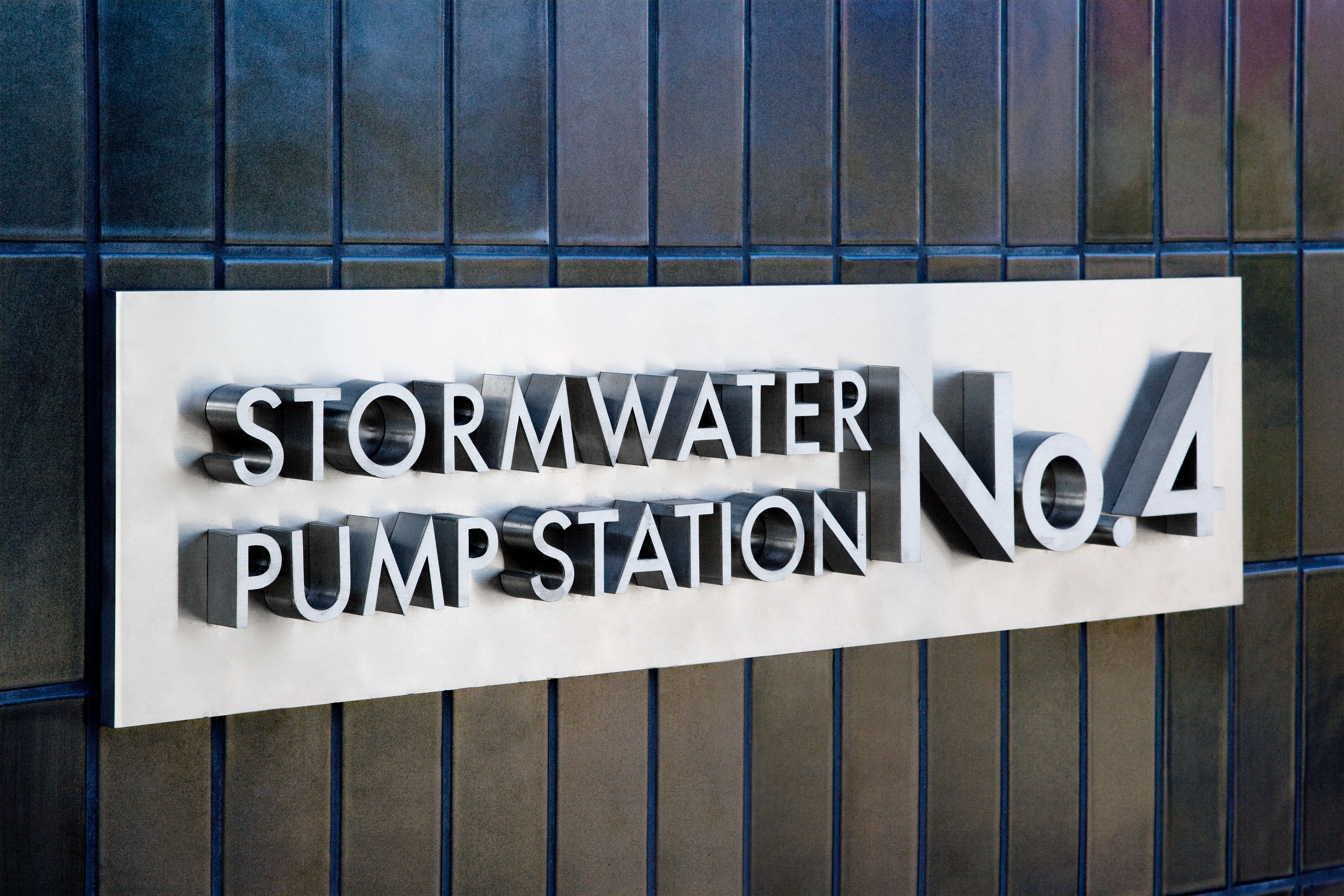 Close-up of a metal sign reading “Stormwater Pump Station No. 4” mounted on a dark tiled exterior wall.