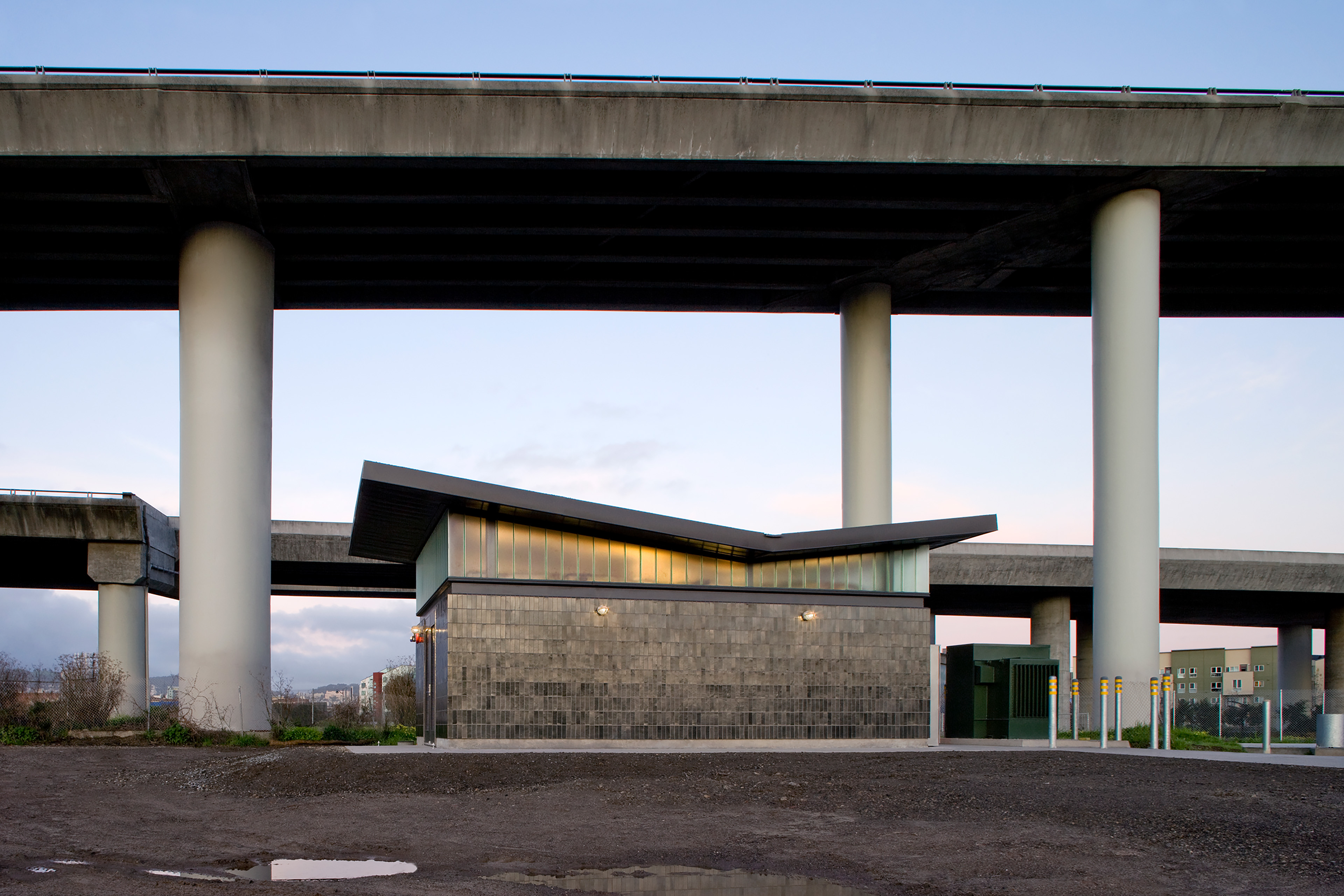 Wide view of a compact utility building beneath intersecting elevated freeways, with large concrete columns and illuminated translucent glazing.