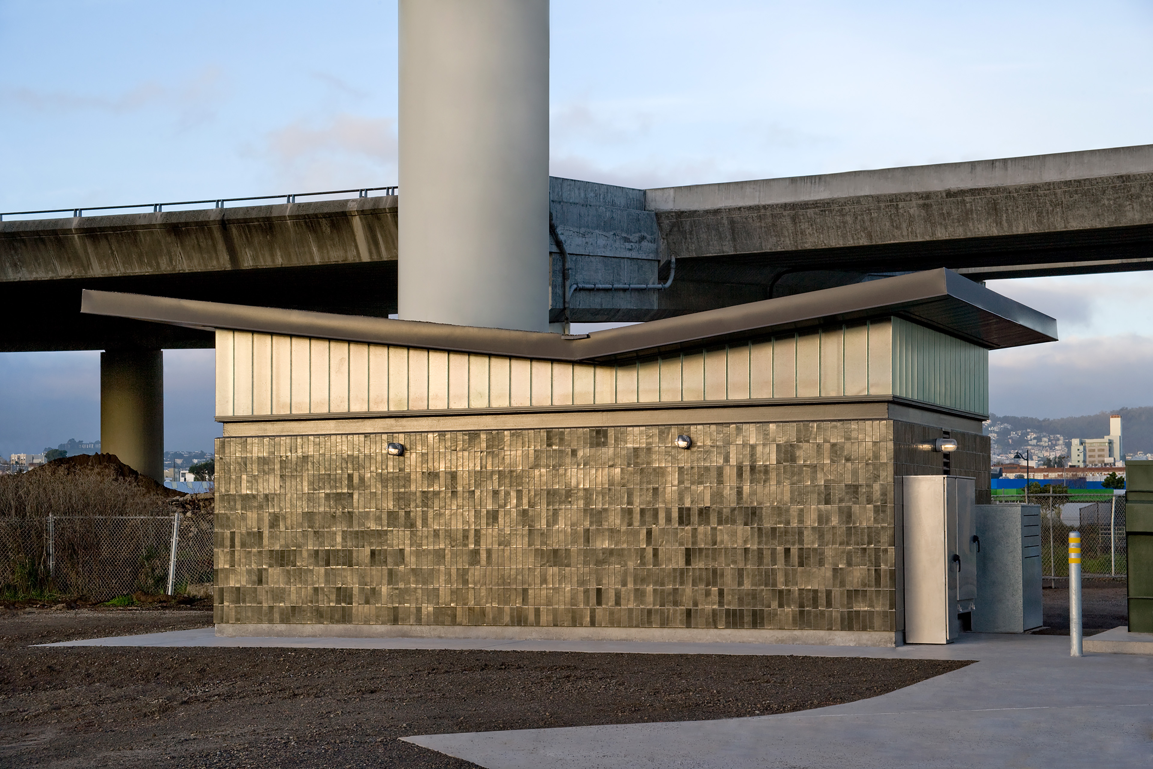Small utility building with gray masonry walls, translucent clerestory glazing, and a folded metal roof, positioned beneath elevated freeway structures.