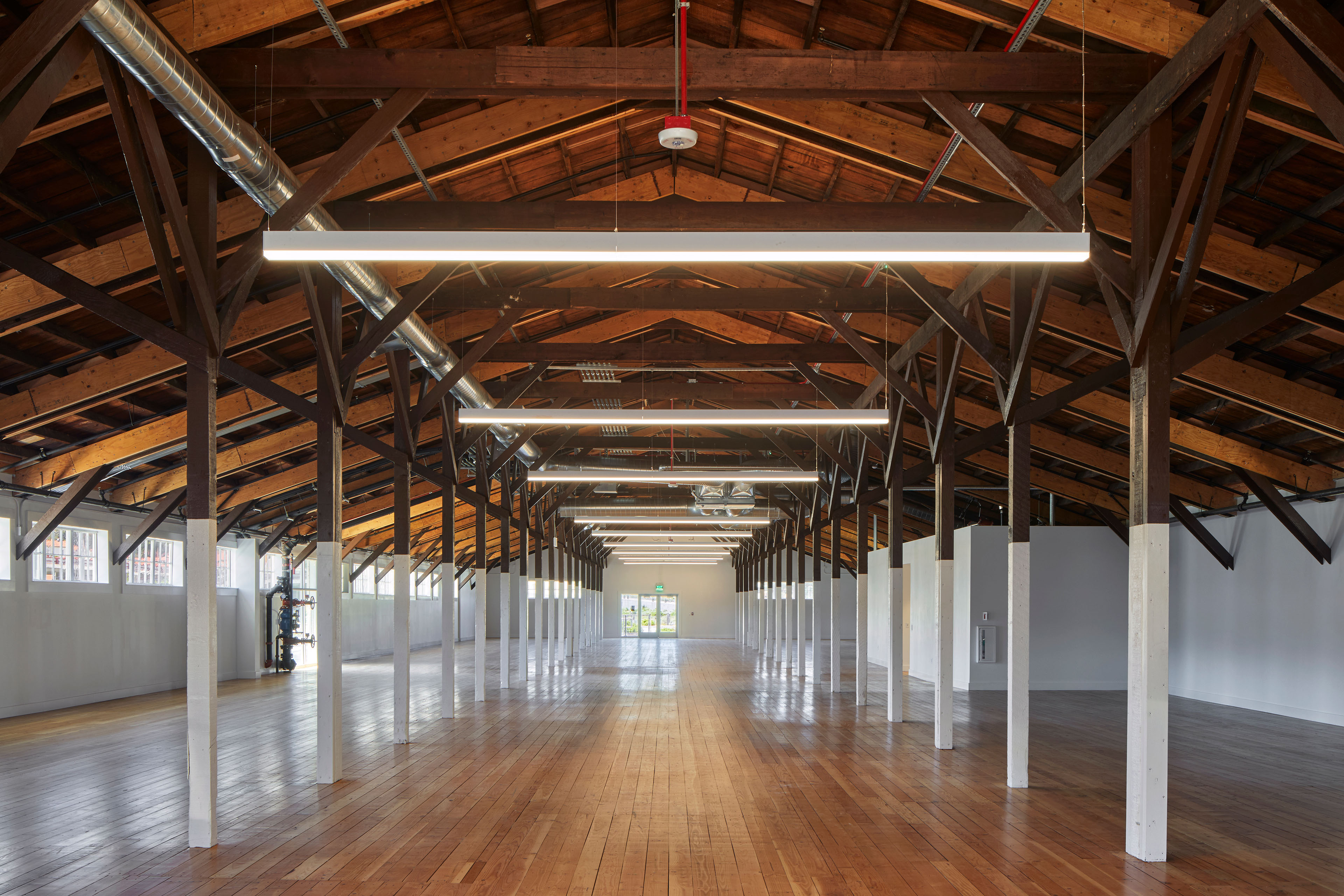Long, open interior with restored wood floors, exposed timber trusses, white columns, suspended linear lights, and visible mechanical ductwork.
