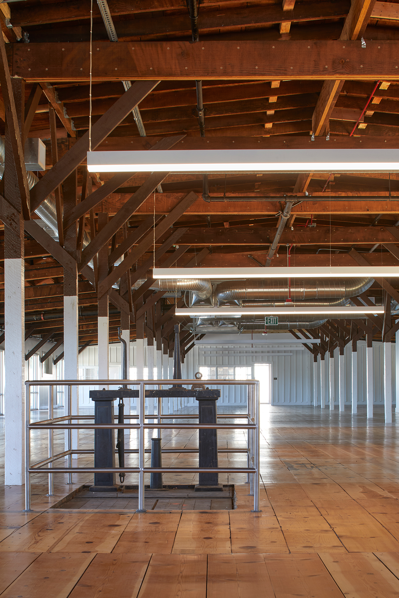 Open historic interior with restored wood floors, exposed timber framing and ductwork, suspended linear lights, and preserved industrial equipment enclosed by a metal railing.