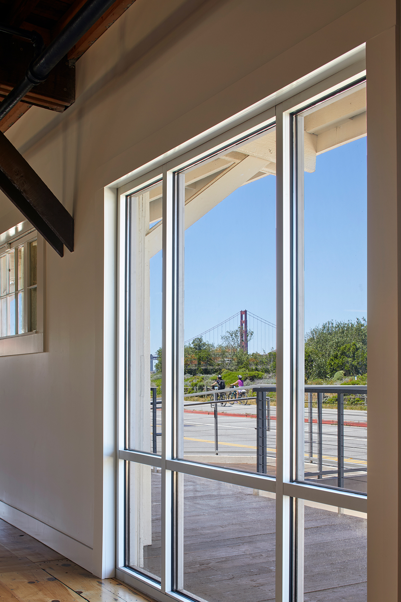 Tall interior windows overlooking an exterior terrace, cyclists, landscaping, and the Golden Gate Bridge.