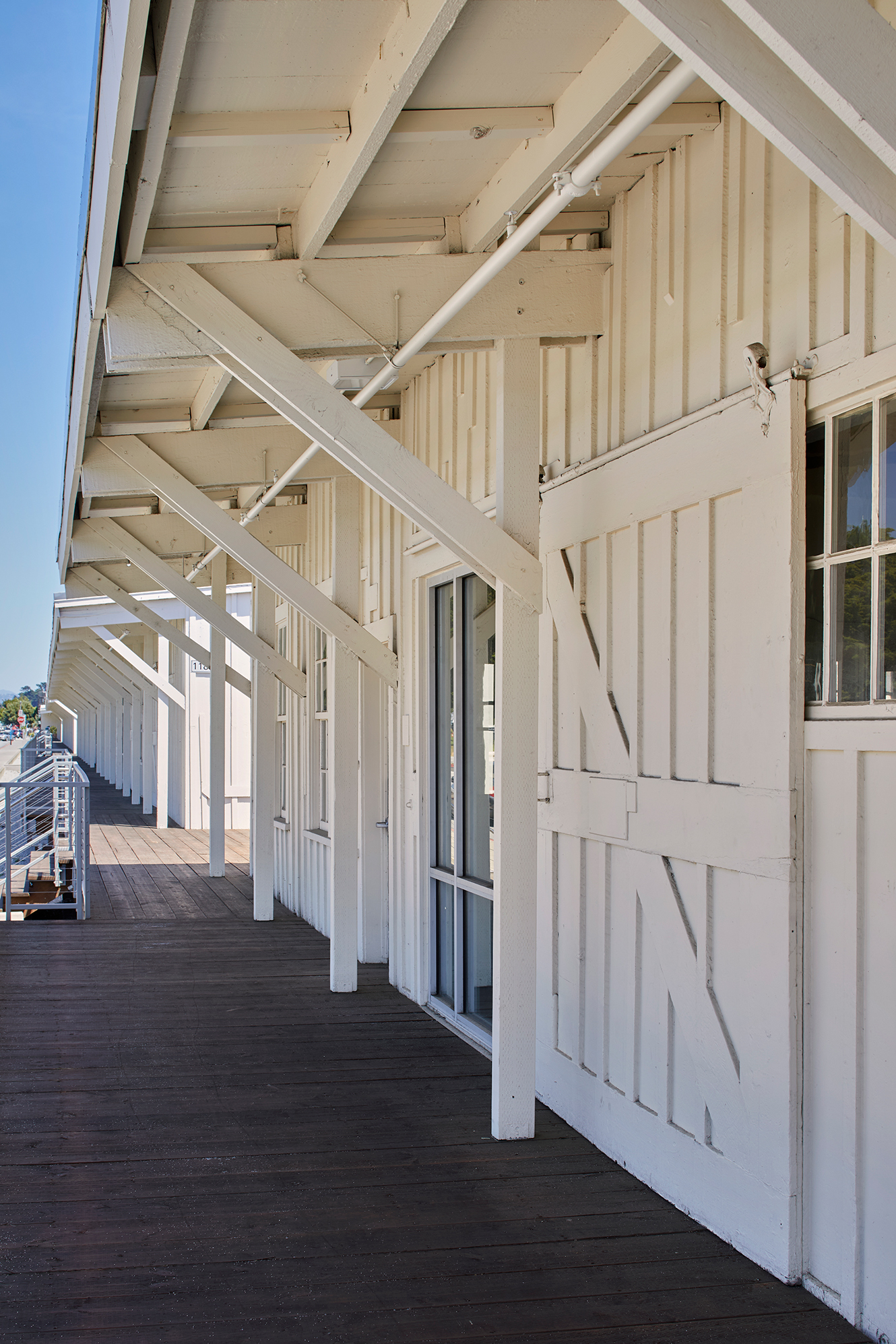 Long covered wooden walkway along a white historic building, with exposed braces, windows, and a metal railing.