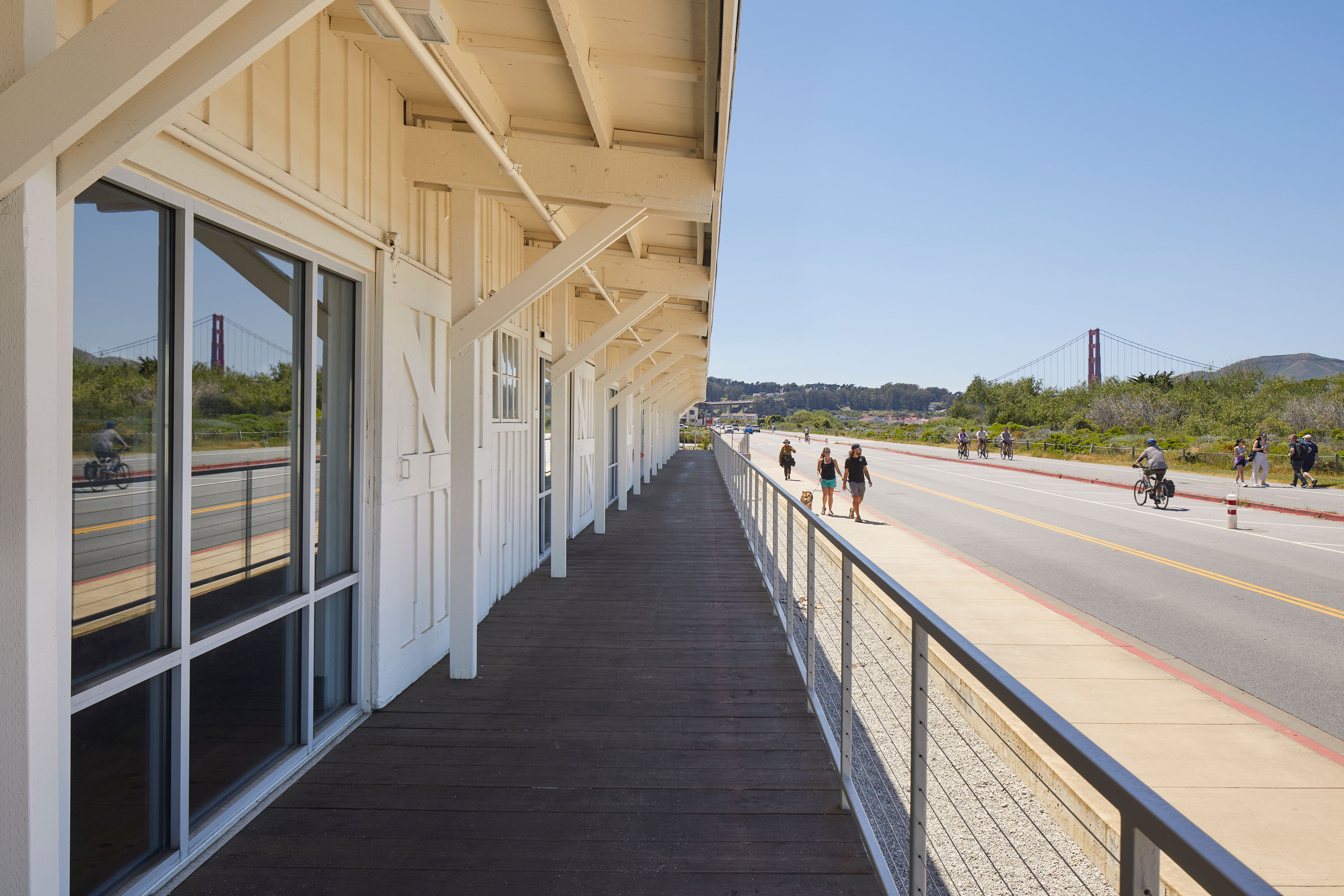 Covered wooden walkway beside a white building, overlooking pedestrians and cyclists on a road with the Golden Gate Bridge in the distance.