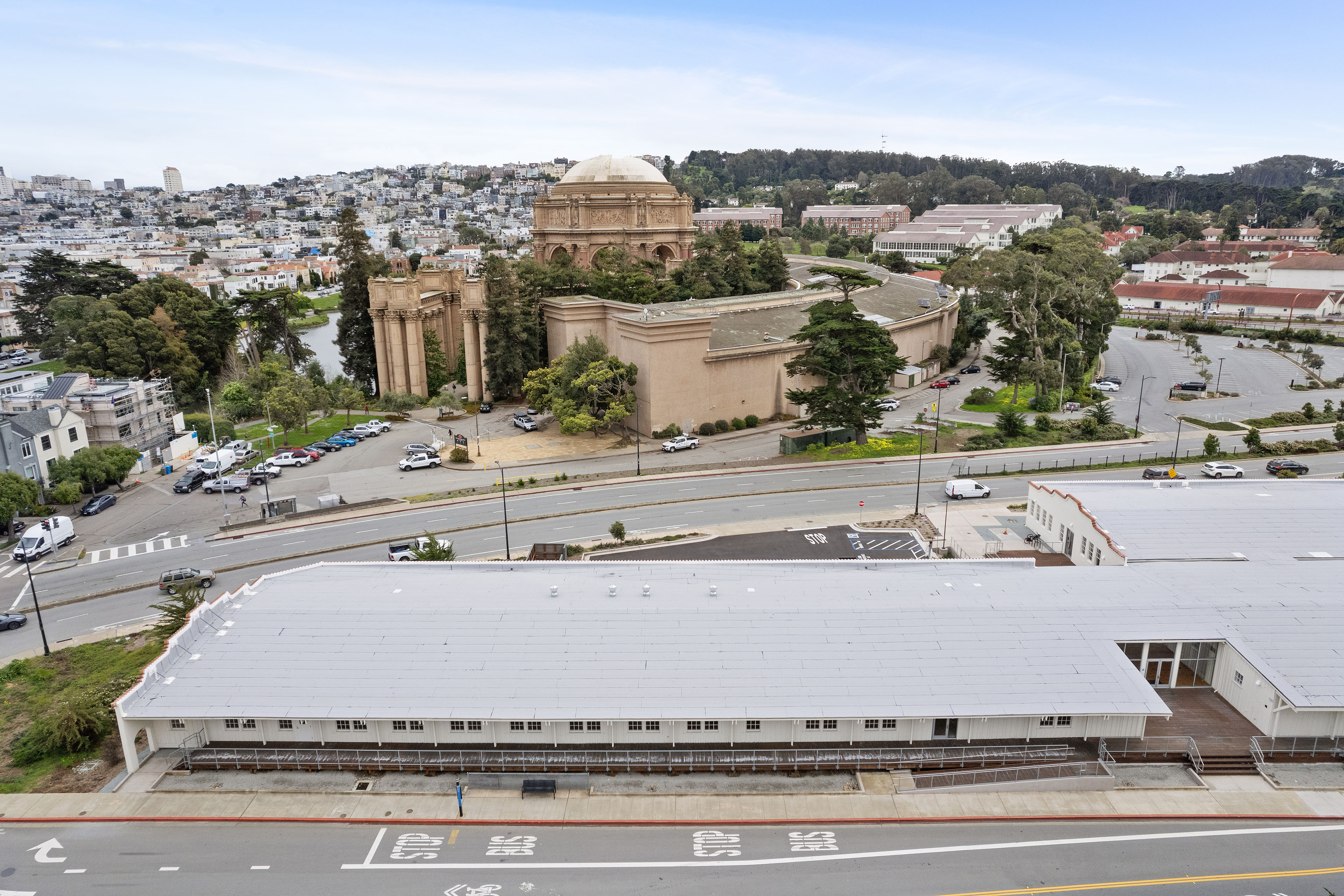Aerial view of a long white historic building across from the Palace of Fine Arts, with surrounding roads, trees, and San Francisco neighborhoods beyond.