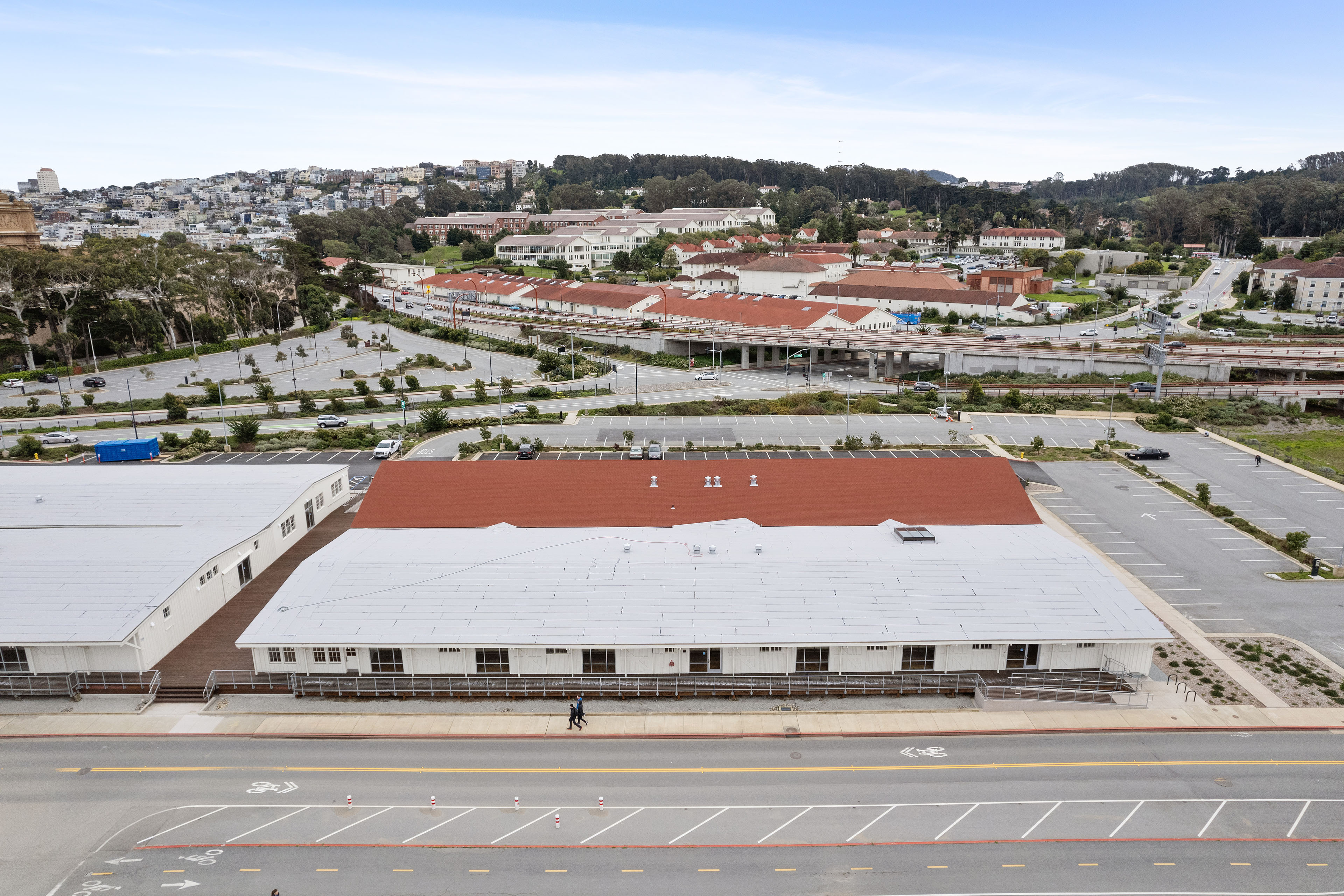 Aerial view of a long white historic building with red and white roofs, bordered by a roadway, parking lots, elevated traffic lanes, and surrounding Presidio buildings.