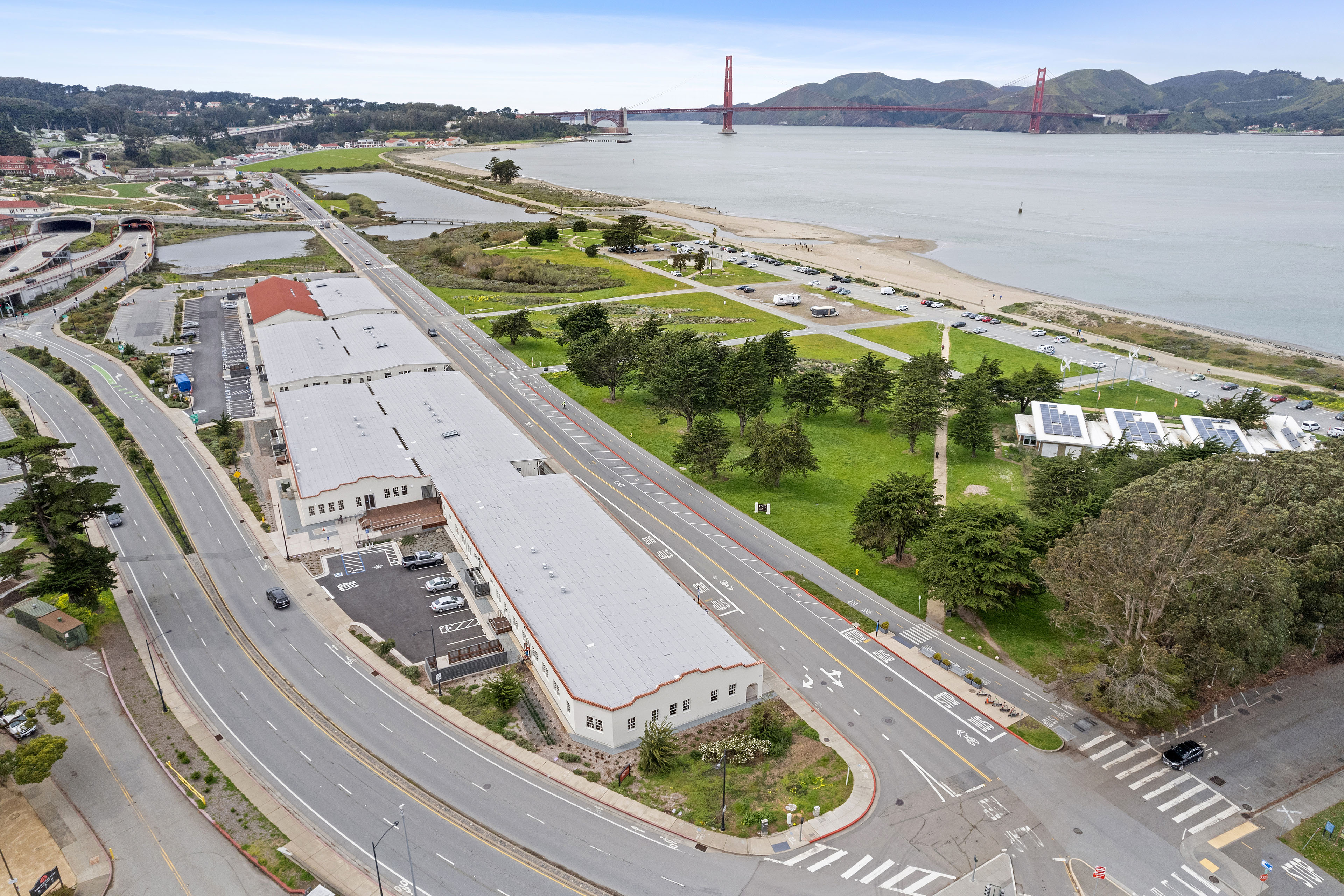 Aerial view of a long historic building beside parkland and the San Francisco Bay, with the Golden Gate Bridge and Marin Headlands in the distance.
