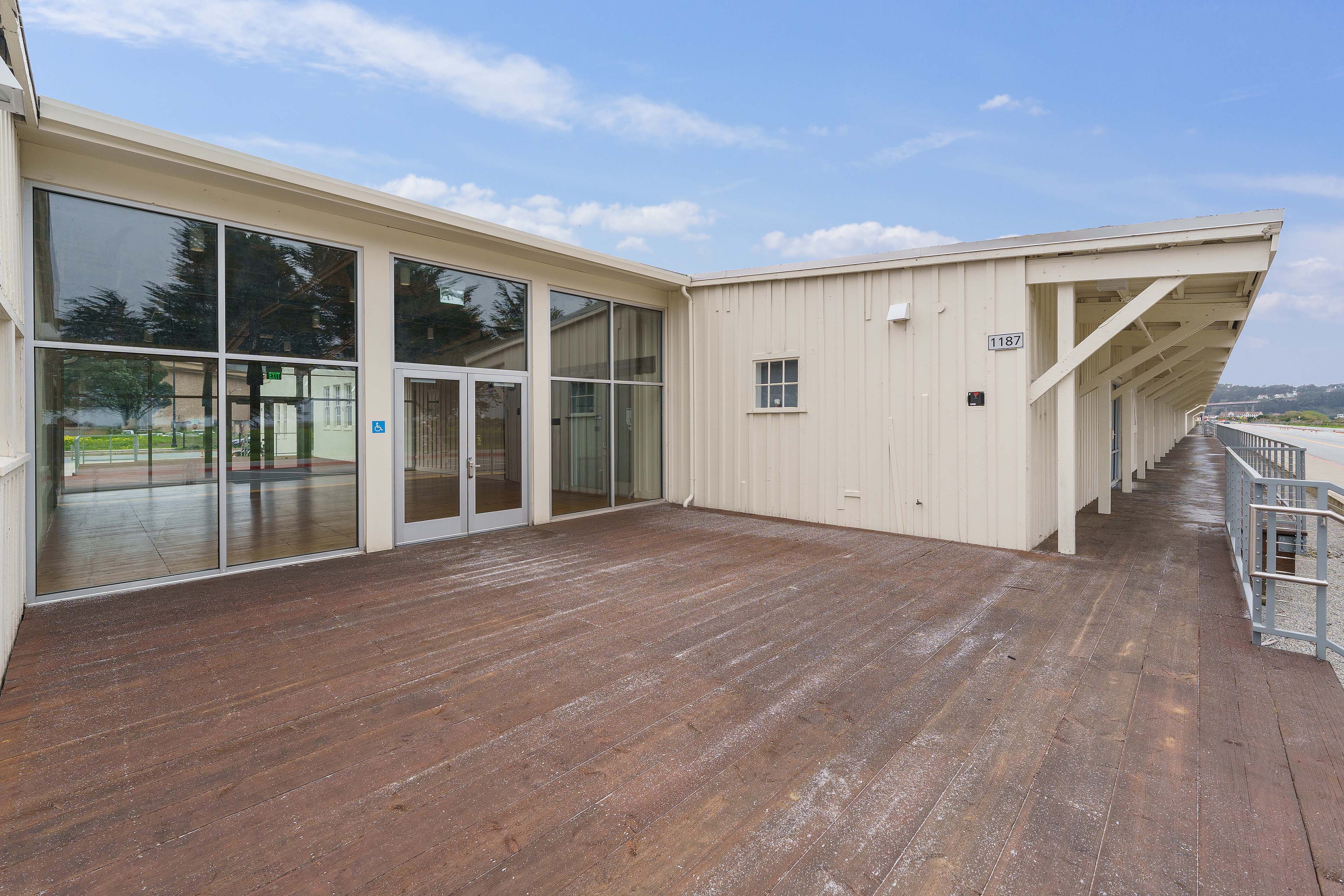 Wooden terrace outside a white historic building, with large glass doors opening to the interior and a covered walkway extending alongside the road.