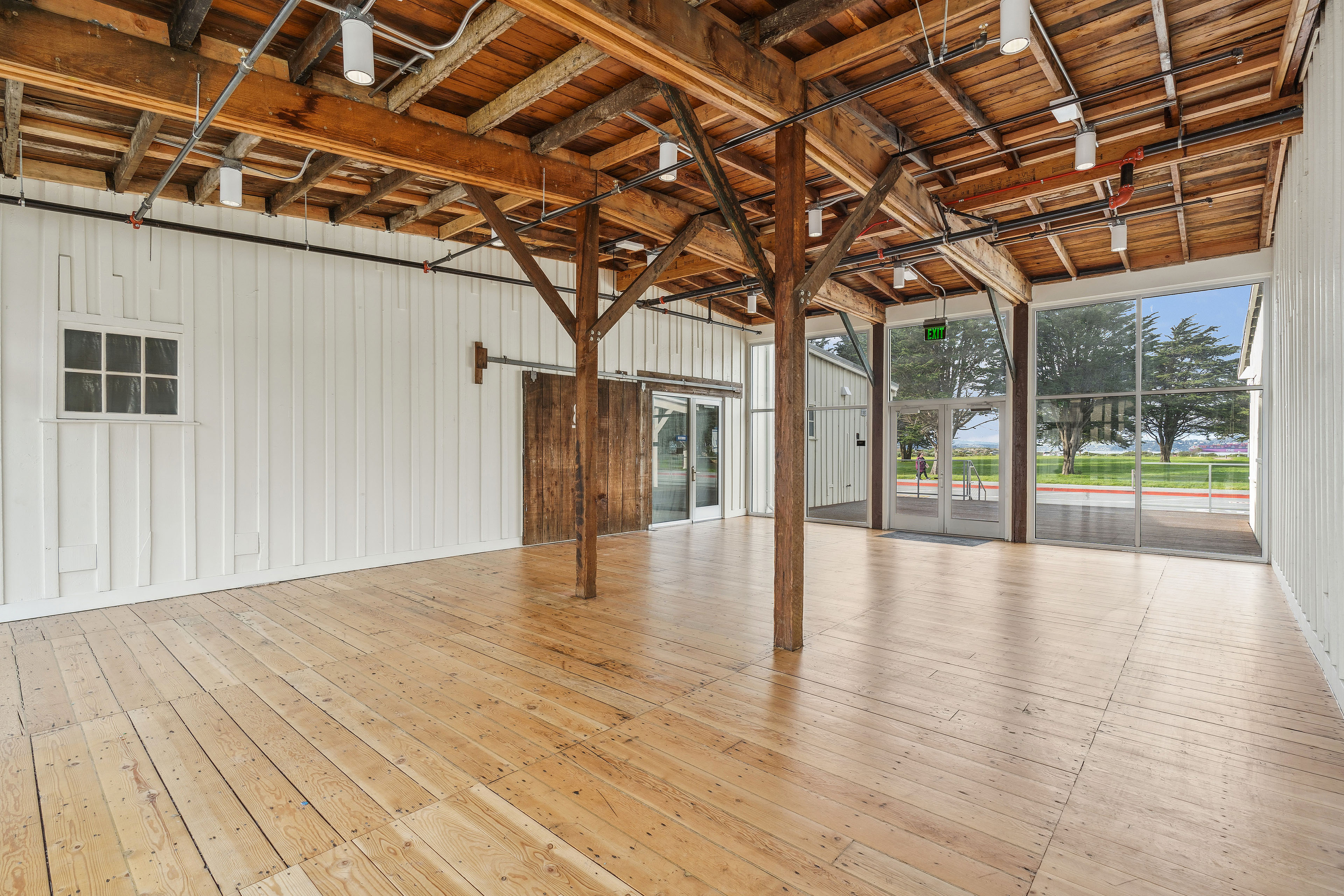 Renovated open interior with exposed timber framing, wood floors, white board-and-batten walls, a sliding barn door, and large glass doors overlooking trees and lawn.