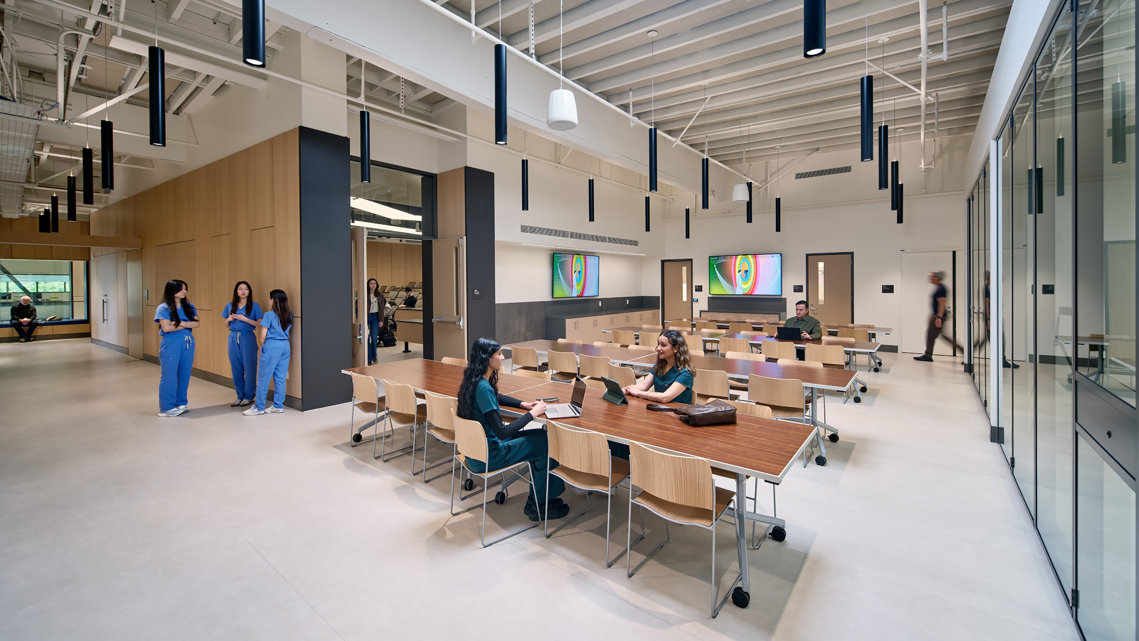 Bright, open learning and collaboration space with movable wood tables and chairs, glass-walled rooms, exposed ceiling structure, and suspended black pendant lights. Several people in medical scrubs gather and work throughout the space, while digital displays and wood-paneled walls define the surrounding teaching areas.