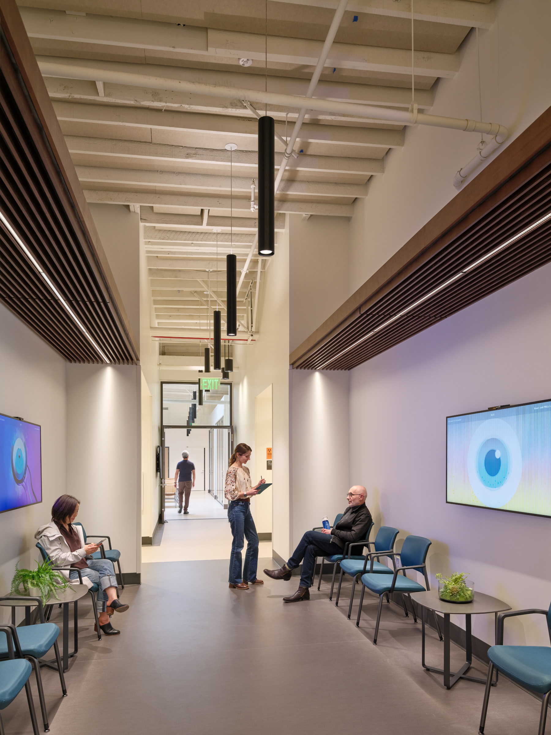 Corridor seating area with blue chairs, side tables, wall-mounted digital displays, exposed ceiling structure, and wood slat accents. Several people sit and converse in the informal waiting space.