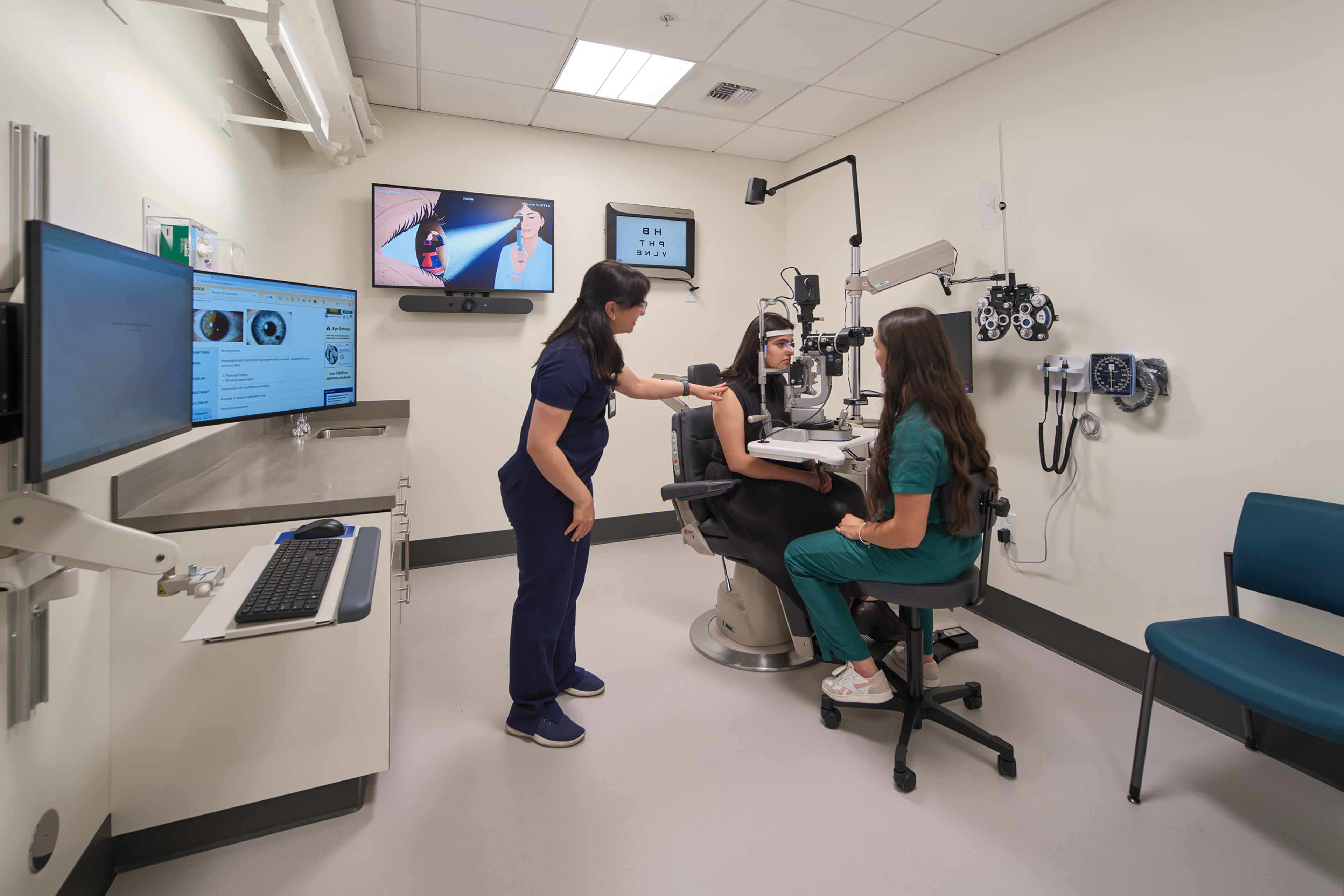 Clinical eye exam room with a patient seated at a slit-lamp microscope while two clinicians assist with the examination. Wall-mounted ophthalmic equipment, computer monitors, and diagnostic displays surround the exam chair.