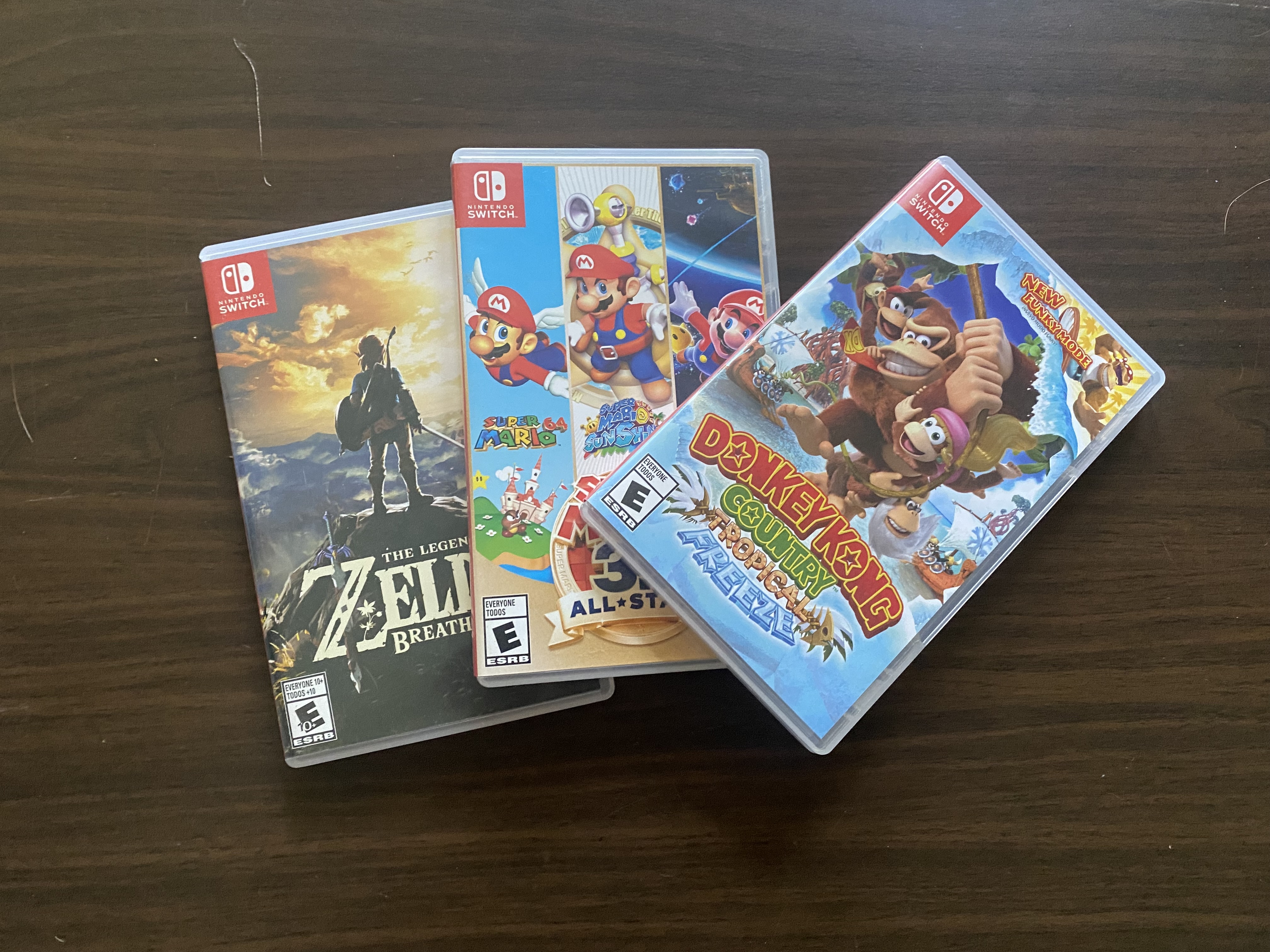 Three Nintendo Switch game cases—The Legend of Zelda: Breath of the Wild, Super Mario 3D All-Stars, and Donkey Kong Country: Tropical Freeze—overlapping on a dark wooden table.