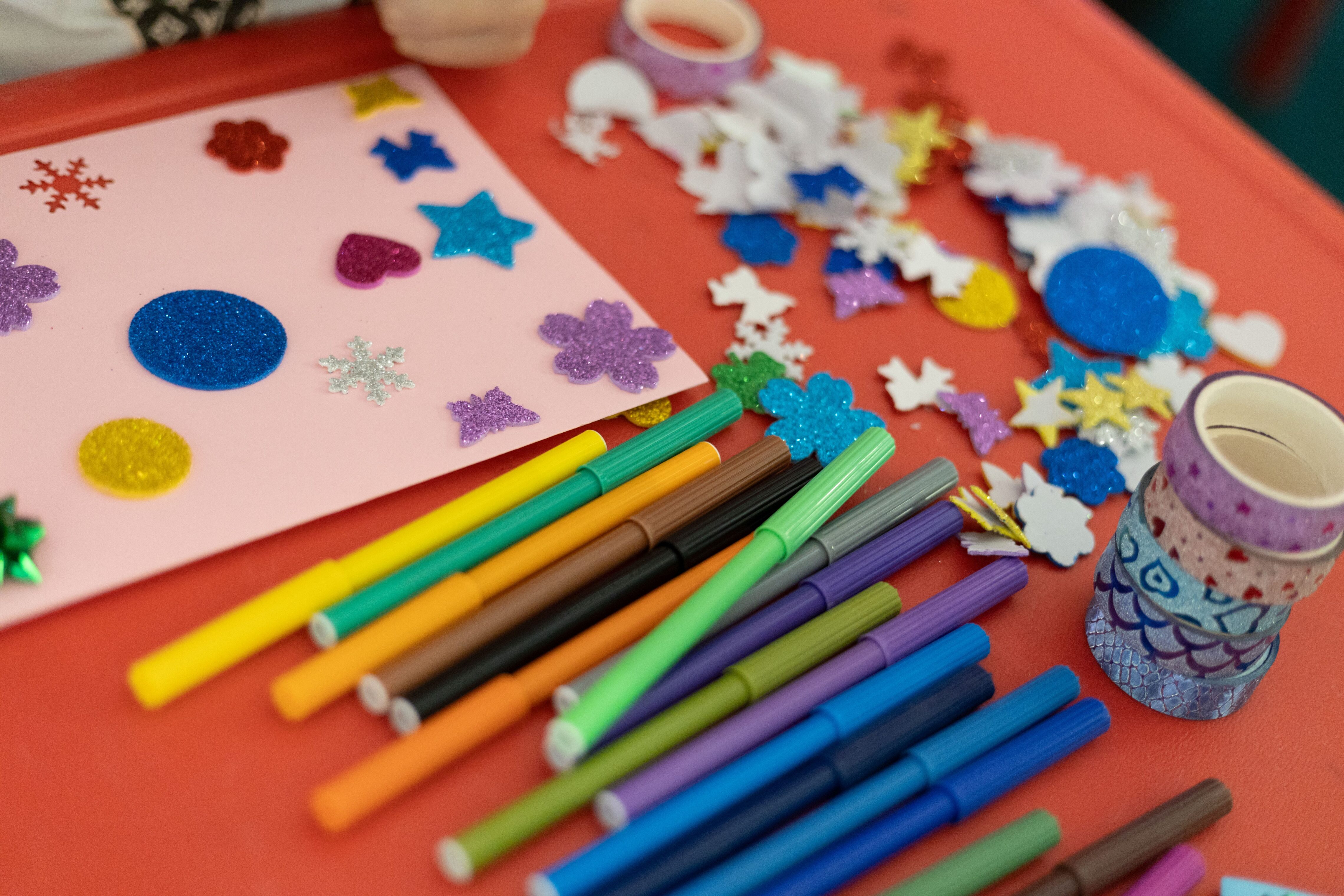 Colorful craft supplies spread across a red table, including markers, rolls of decorative tape, glittery foam shapes, and a pink sheet decorated with stars, hearts, snowflakes, and other shapes.
