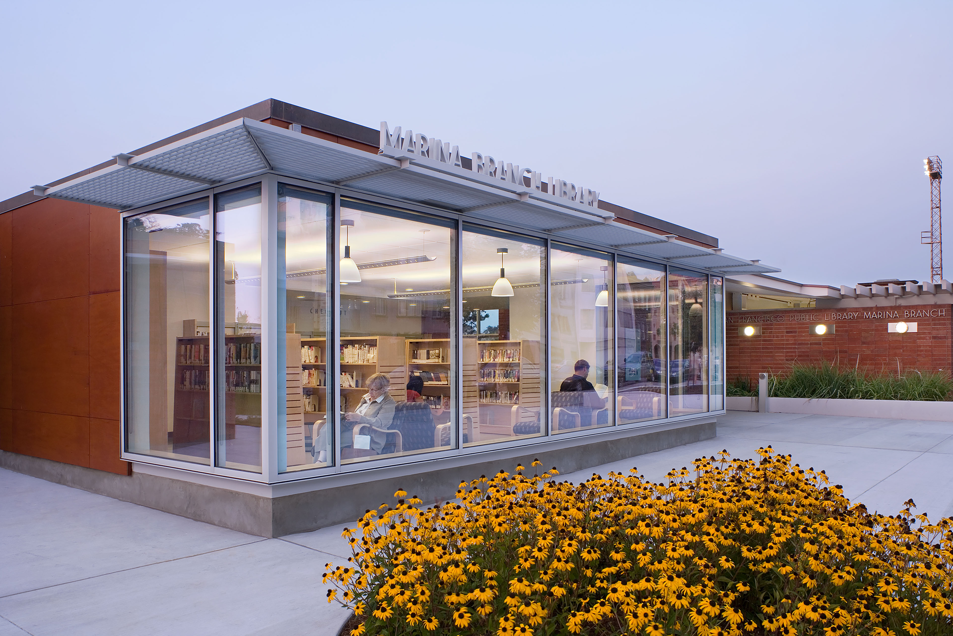 The modern, low-profile exterior of the San Francisco Public Library Marina Branch captured at dusk. The building features an expansive glass-walled pavilion with warm indoor lighting that showcases bookshelves and patrons reading inside. Large, metal signage reading 