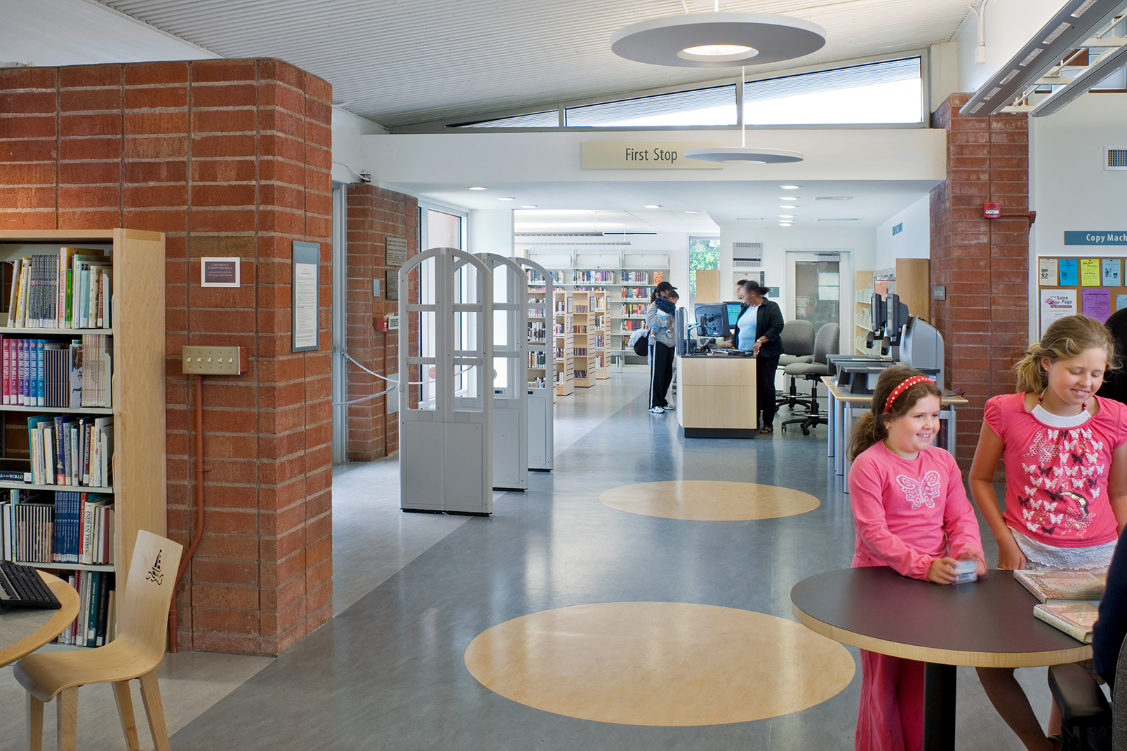Interior view of a brightly lit, modern public library branch. On the left, a large brick column stands next to a wooden bookshelf filled with books. In the foreground, two young girls sit at a round table, one wearing a pink shirt with a butterfly graphic. In the background, patrons stand near a service desk under a sign reading 
