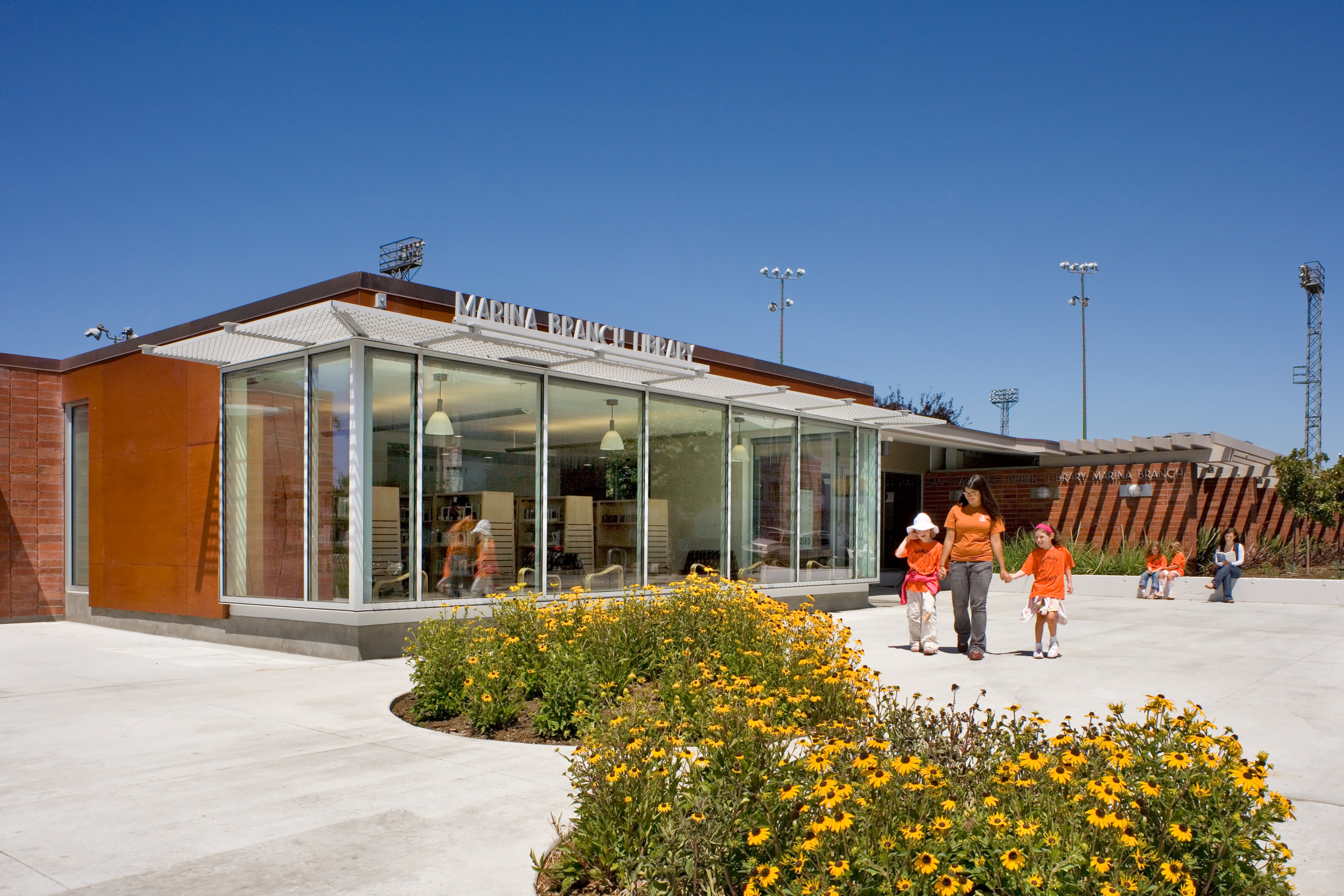 Exterior of the Marina Branch Library on a sunny day, featuring a glass-walled reading room, landscaped courtyard with yellow flowers, and families walking near the entrance.
