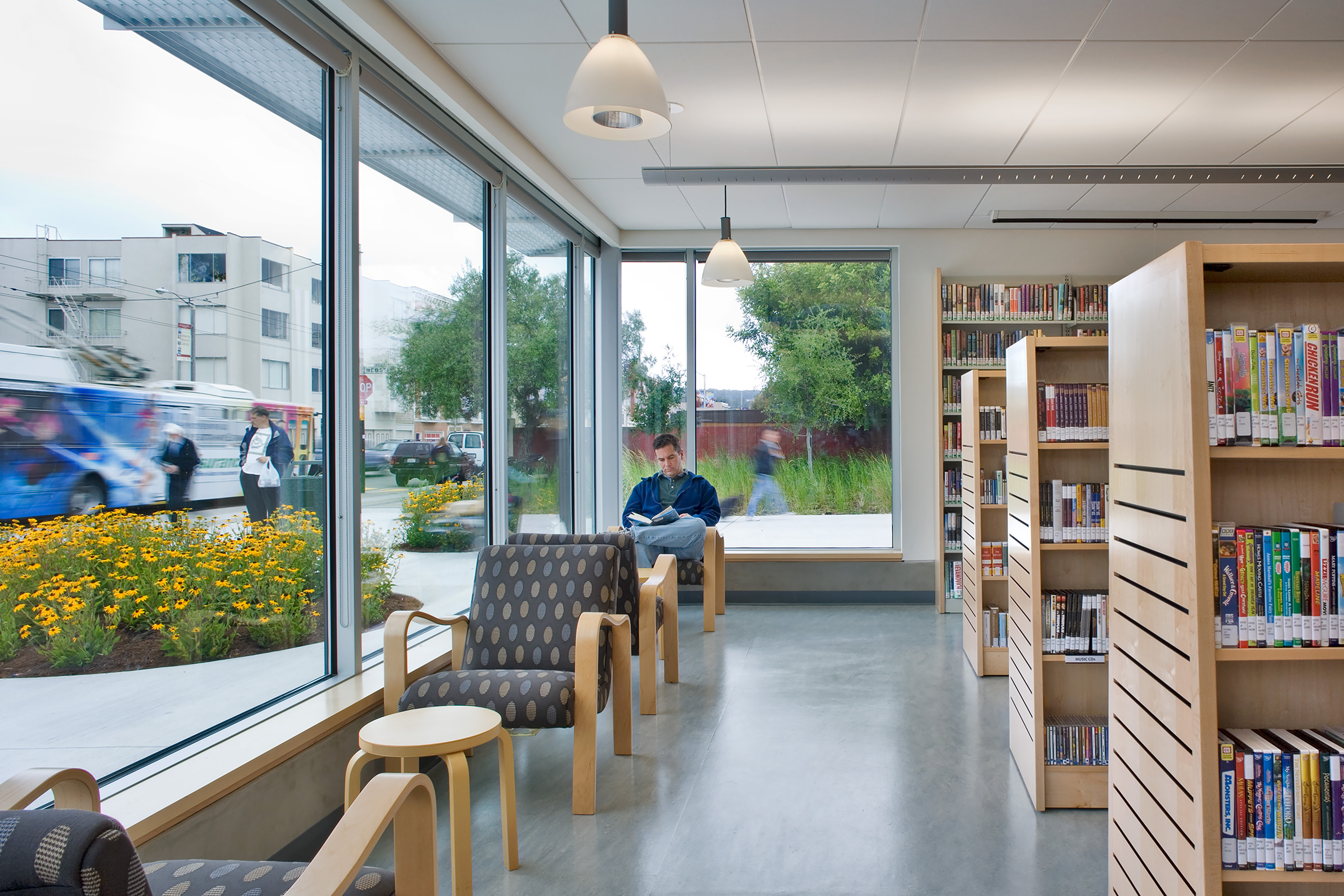 A brightly lit, modern public library interior featuring a seating area with pattern-upholstered armchairs and wooden accent tables positioned along a large expanse of floor-to-ceiling glass windows. Outside the window, vibrant yellow flowers bloom in a landscaped planter, and a city bus with graphic wraps drives down a city street. To the right, neat rows of light-wood bookshelves hold media materials and books. A patron sits quietly in an armchair reading a book near the far window, under sleek hanging pendant light fixtures.