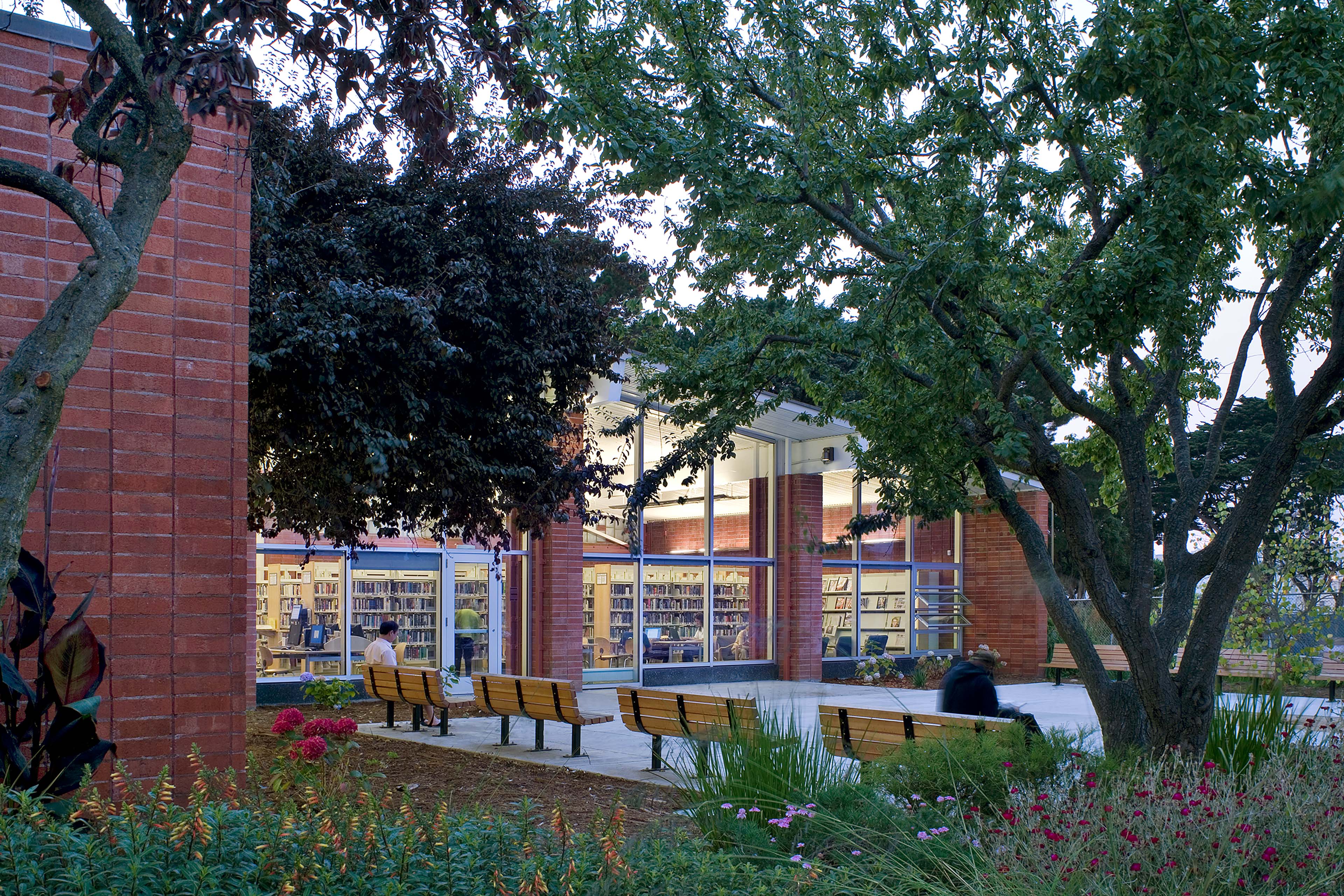 An exterior twilight view of the library building's rear facade, constructed with warm red-brick columns and soaring, floor-to-ceiling glass windows that glow from interior lights. A concrete courtyard in front of the windows is furnished with several wood-slat benches arranged beneath mature, leafy trees. The entire scene is framed by lush, landscaped garden beds filled with pink and red wildflowers and green ornamental grasses.