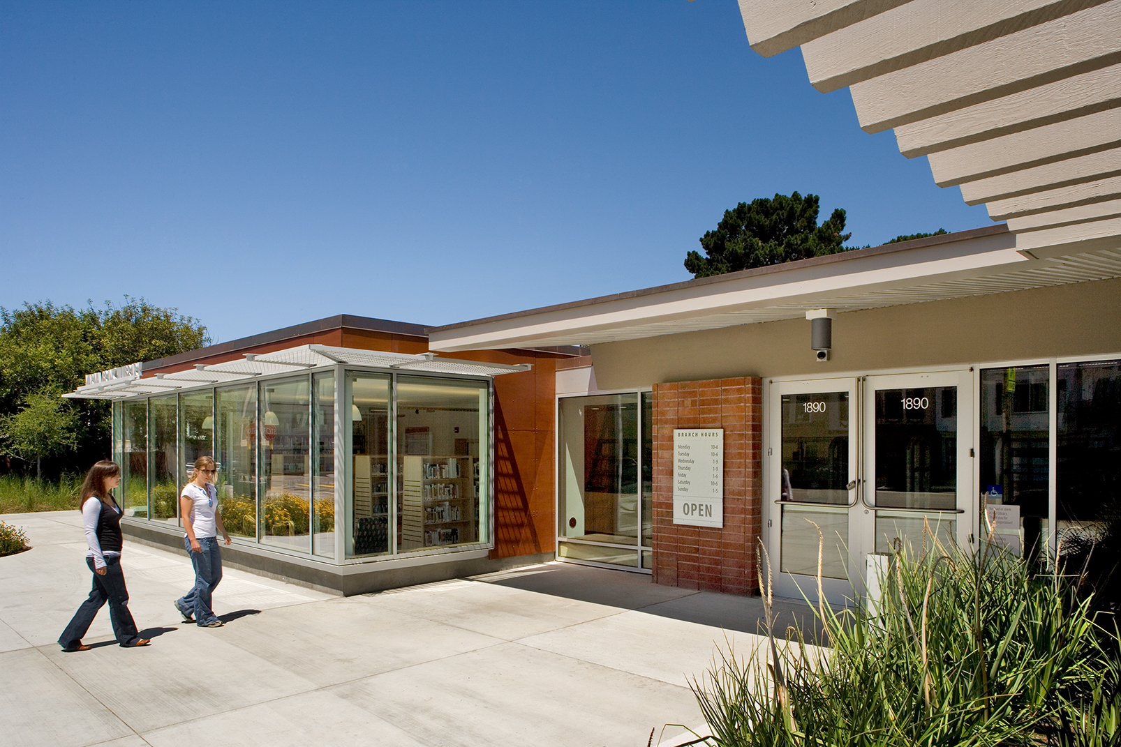A daytime exterior view of a modern, single-story public library building with large glass windows showing the bookshelves inside. Two women are walking along a concrete pathway in front of the building. The entrance doors display the address number 1890, and a framed sign next to the door lists the branch hours under a clear blue sky.