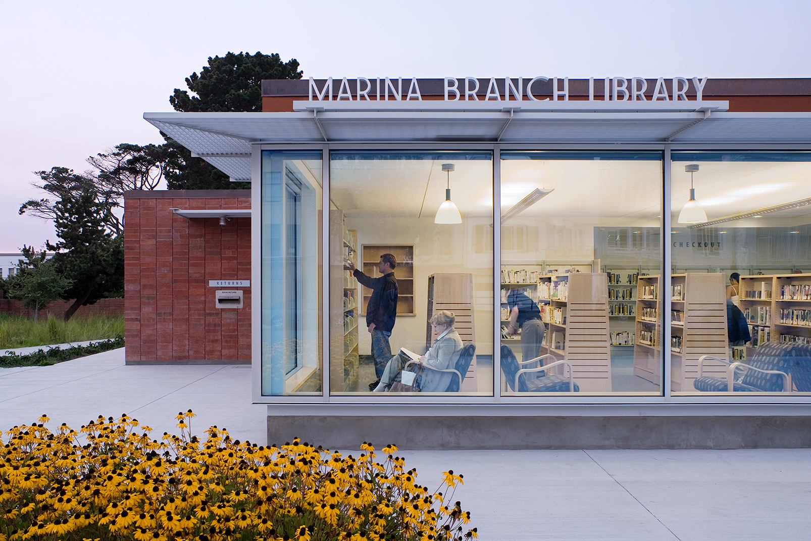 An evening view of the exterior of the Marina Branch Library, highlighting its large glass facade glowing with warm interior light. Inside, people can be seen browsing bookshelves and sitting in armchairs reading. In the foreground, a vibrant bed of yellow black-eyed Susan flowers blooms along the walkway under a twilight sky.