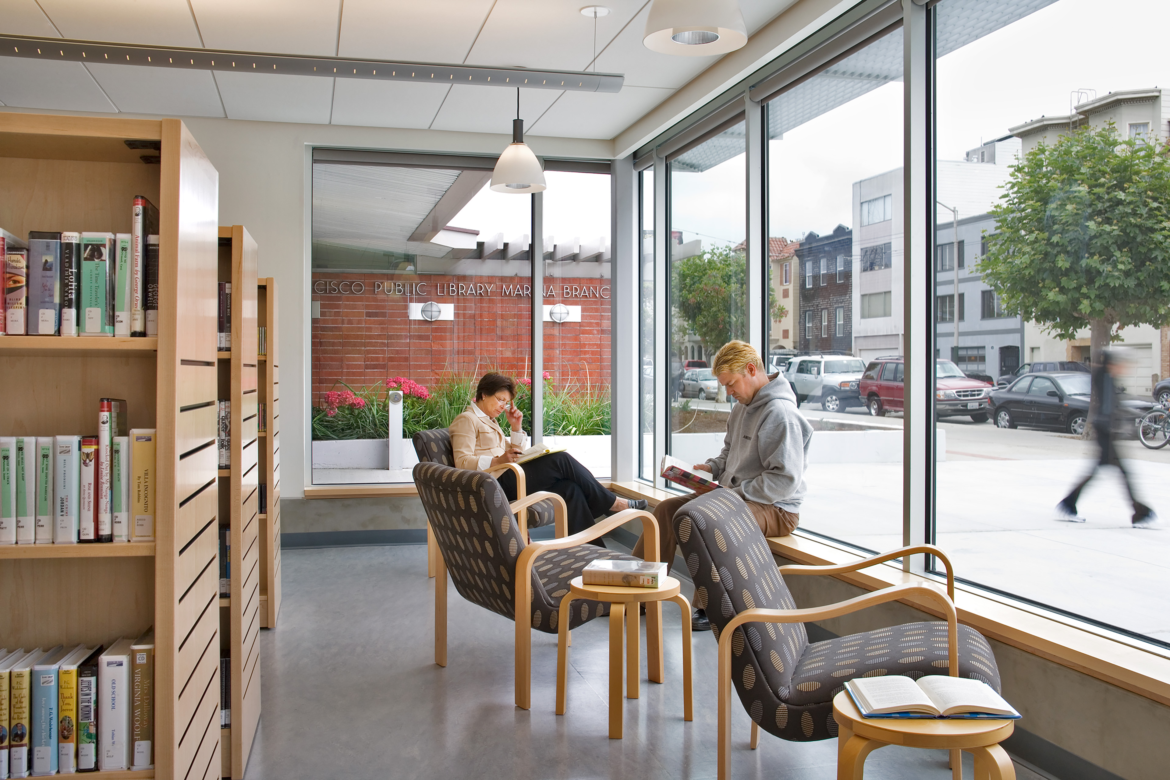 An alternate angle inside the modern public library highlights a light-filled reading corner next to extensive floor-to-ceiling windows overlooking a street with parked cars and storefronts. Two library patrons sit across from each other in comfortable, brown pattern-upholstered armchairs while reading books. Light-colored wooden bookshelves packed with books stand to the left, and a low wooden accent table sits between the chairs with a book resting open on it. Through the window glass, a brick building facade is visible, displaying signage that reads 