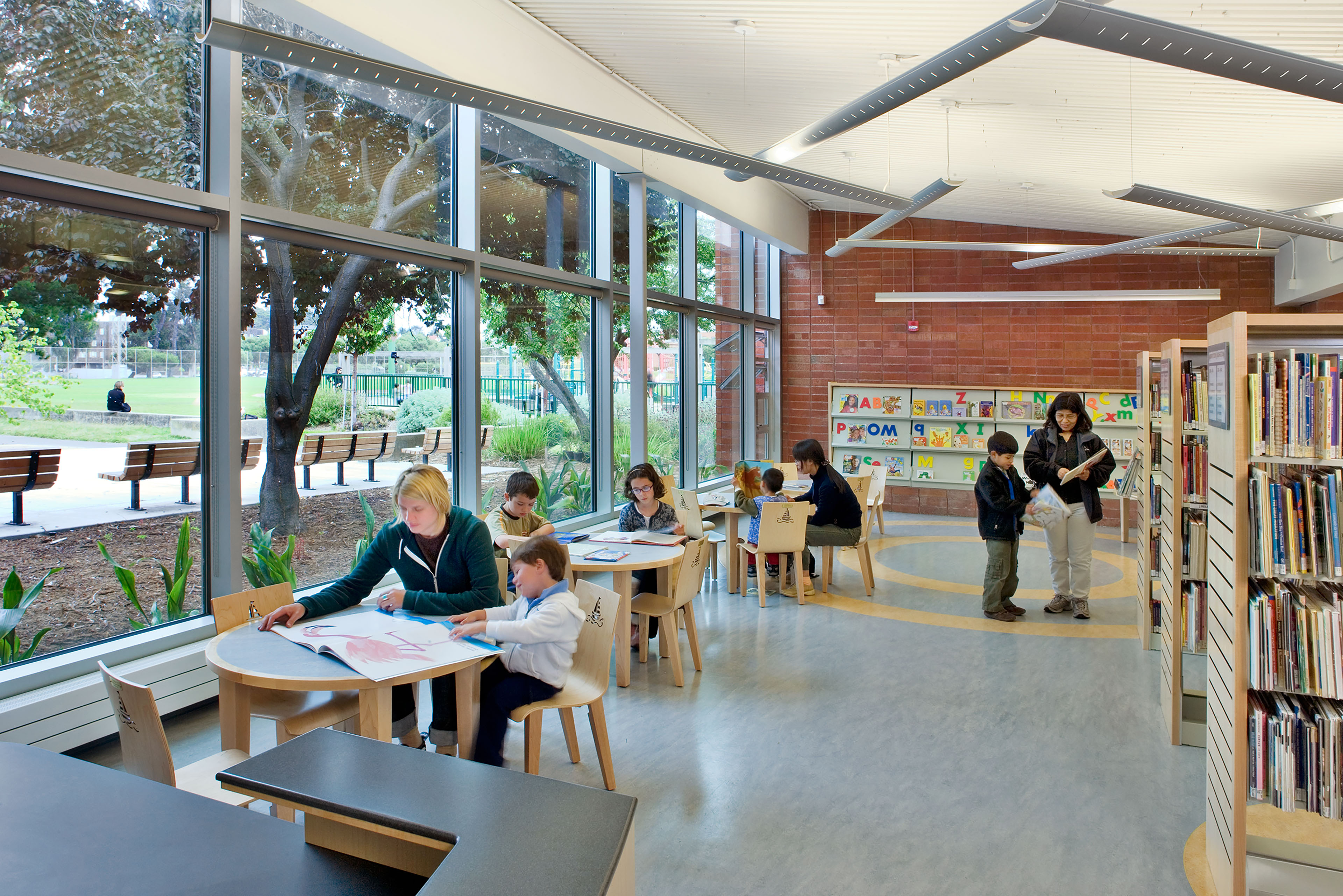A bright and inviting reading area tailored for children within a library, flanked by tall glass windows looking out onto a park with green grass, trees, and benches. Several small, child-sized wooden tables and chairs are arranged along the window wall where children and parents sit together looking at books. To the right, tall wooden book stacks run parallel to the space, and a colorful alphabet display board is visible on the far brick wall in the background.