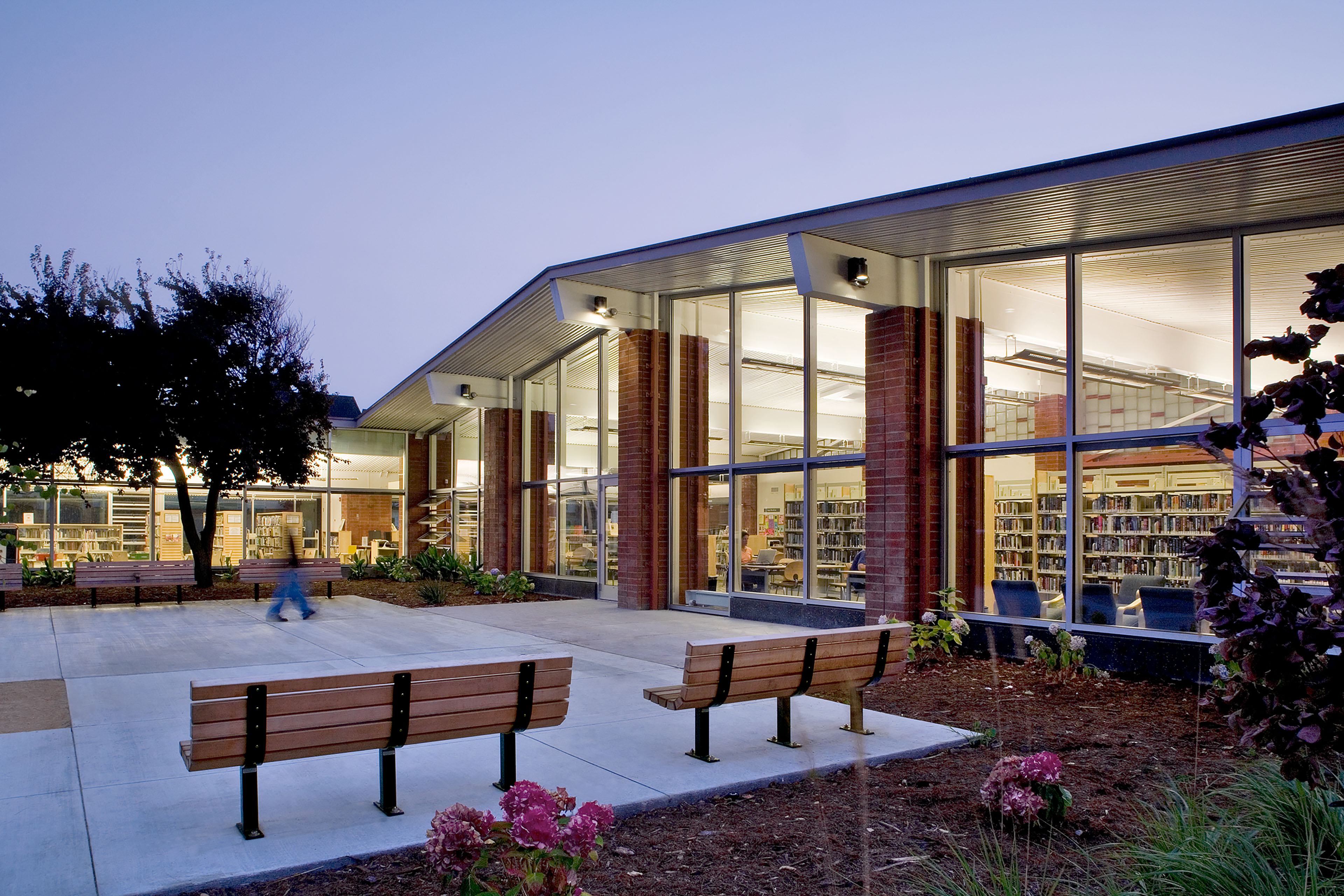 Exterior courtyard of the Marina Branch Library at dusk, with benches, landscaping, and floor-to-ceiling windows revealing the warmly lit reading room and bookshelves inside.
