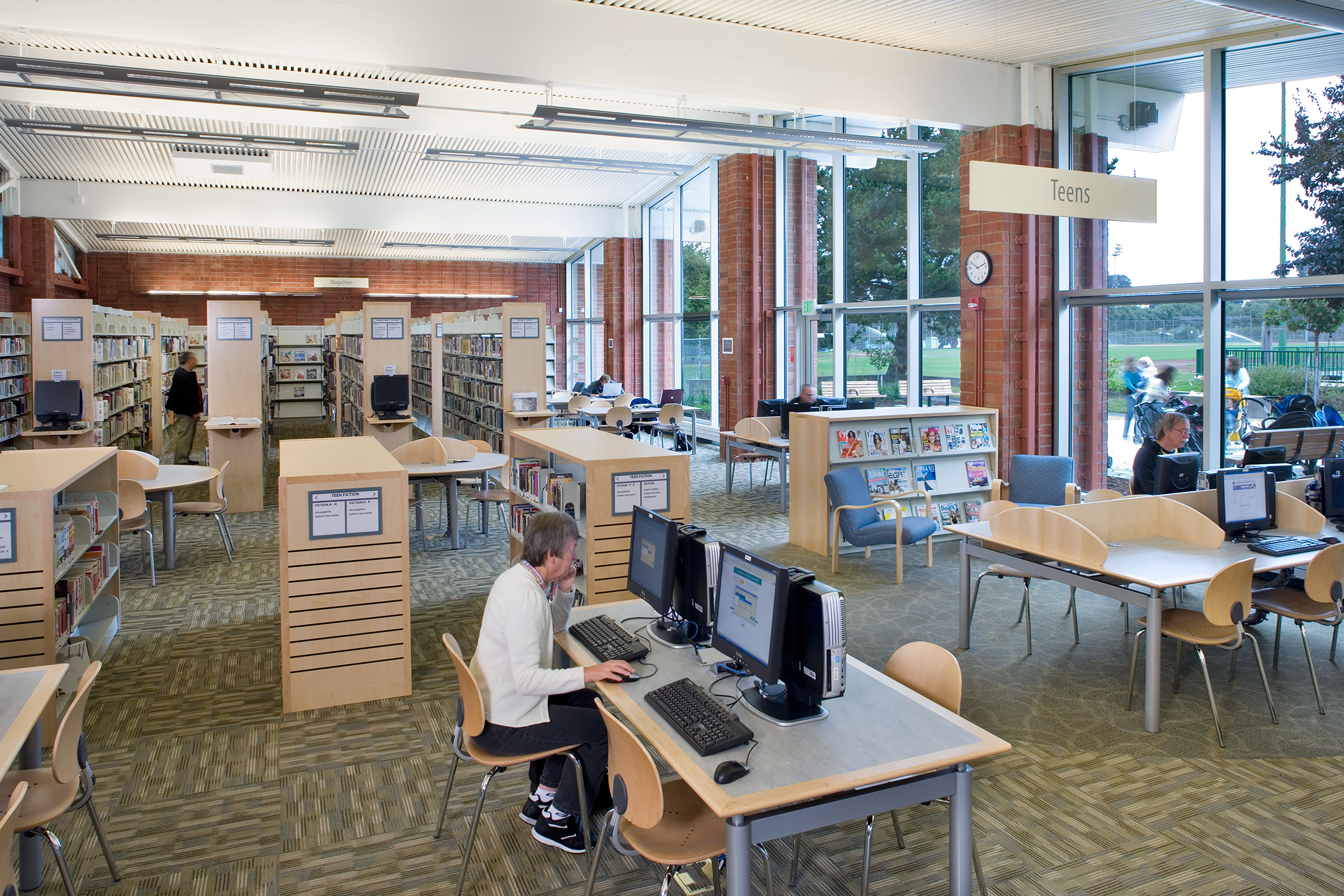 A spacious, wide-angle interior view of a modern public library branch. In the foreground, a patron sits at a rectangular table utilizing a desktop computer station. The open-plan layout features pattern-carpeted flooring, multiple rows of light-wood bookshelves, circular study tables, and additional computer hubs. Massive floor-to-ceiling glass windows flank the right wall, exposing an outdoor park area and filling the space with natural light. A prominent hanging sign near a brick structural pillar marks the 