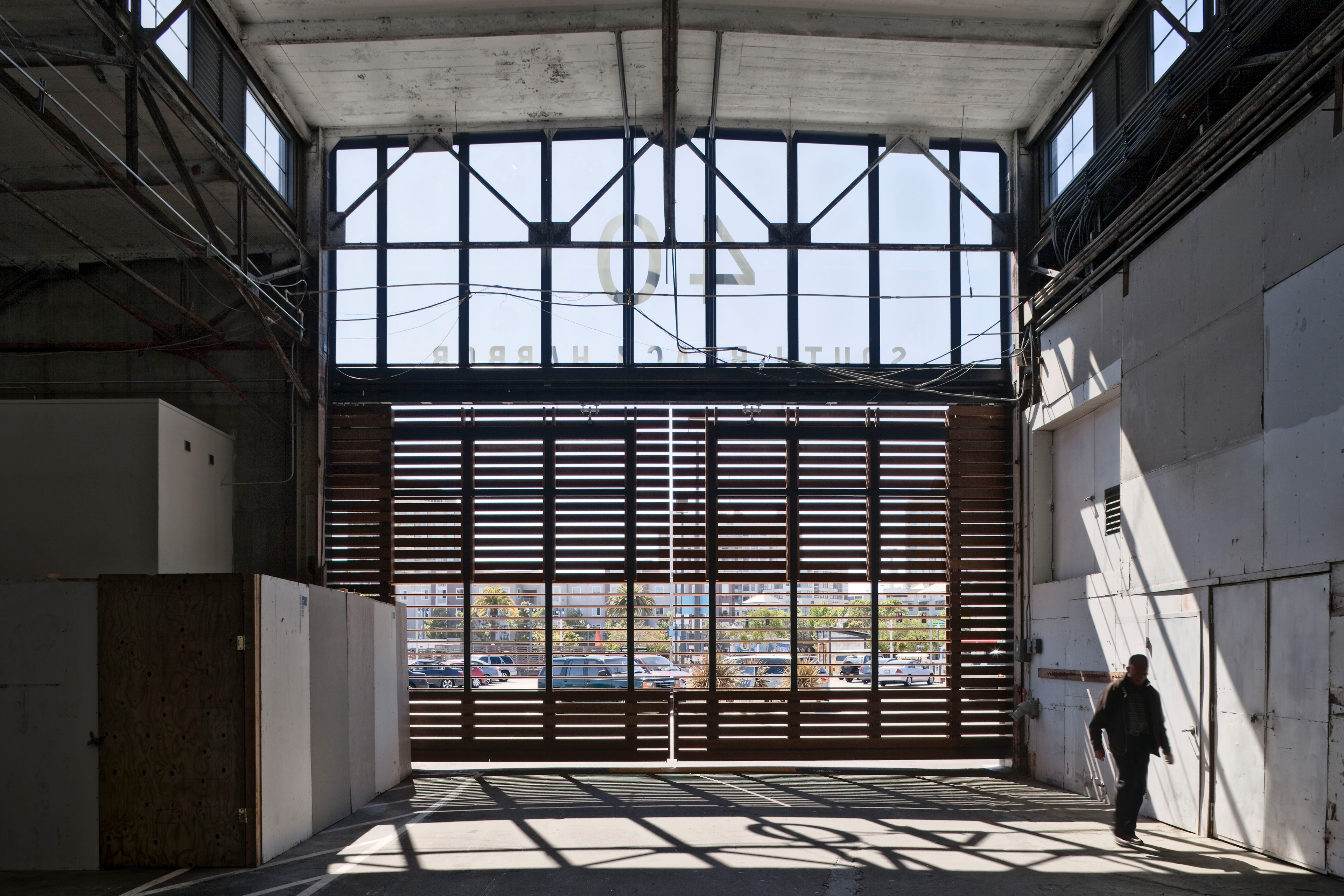 Interior of a renovated harbor building with exposed steel framing, high clerestory windows, and large wood-slat doors filtering daylight into the space.