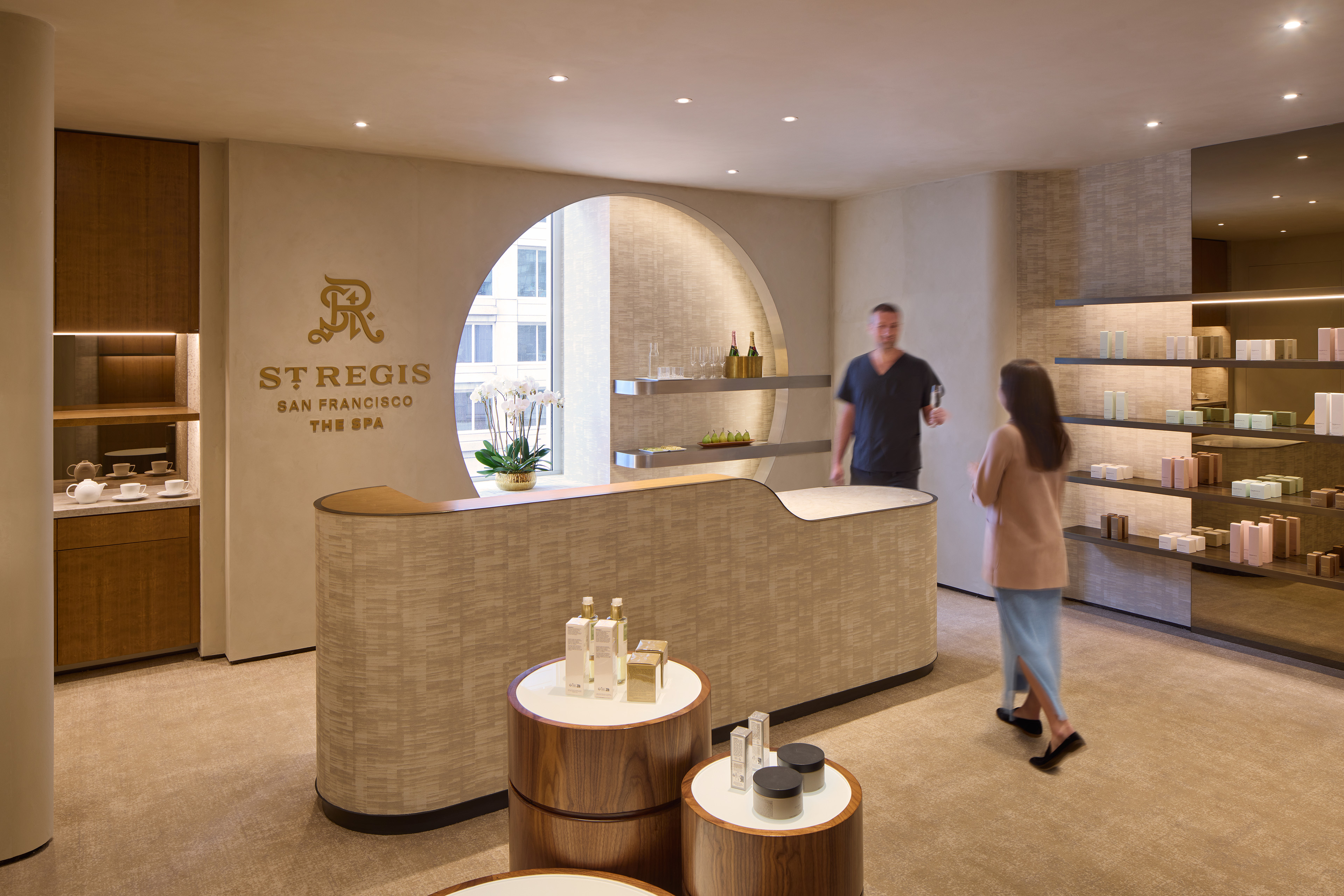 Reception area of The Spa at The St. Regis San Francisco, featuring a curved desk, arched opening, warm wood accents, textured finishes, product displays, and two people standing near the desk.