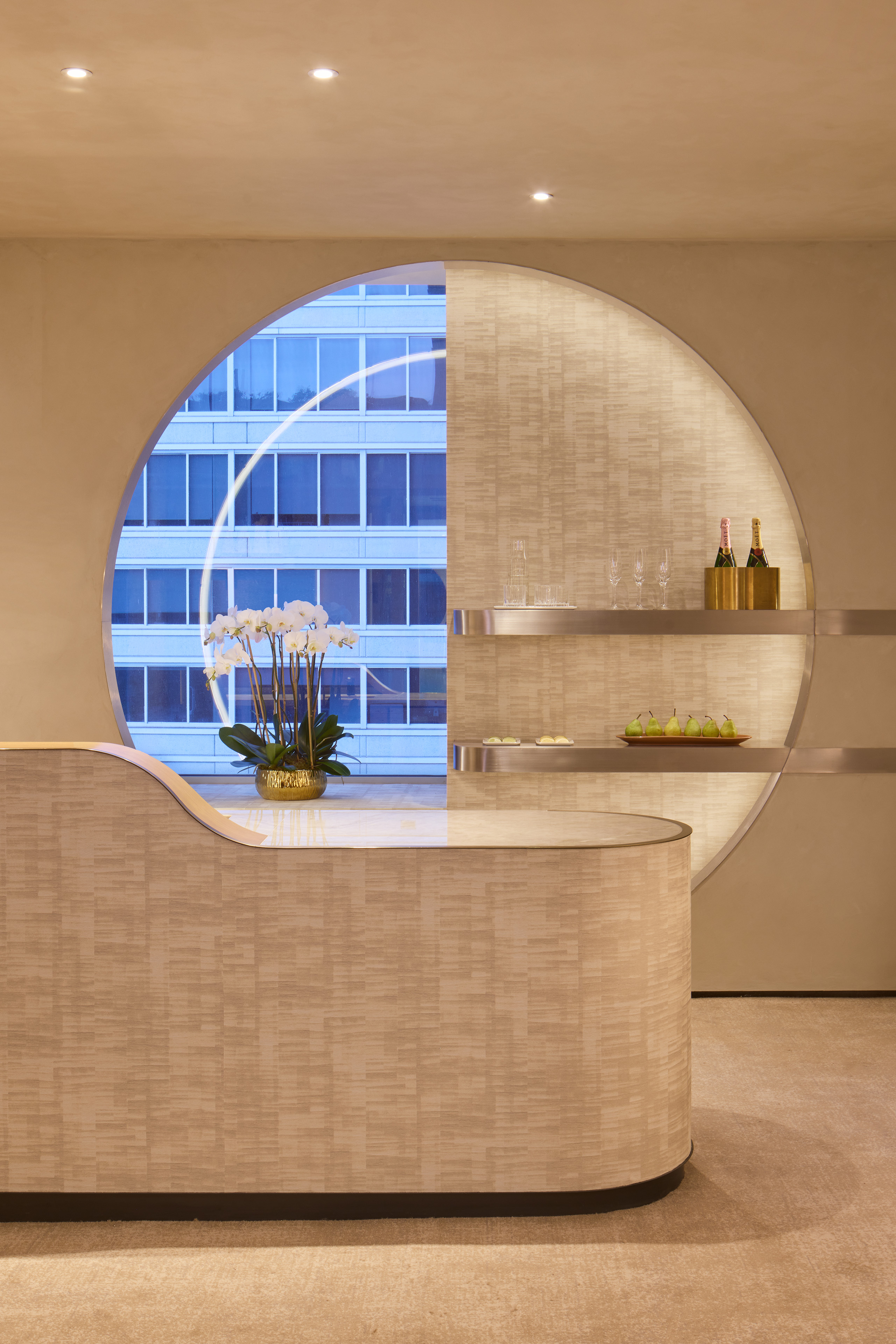 Reception desk at The Spa at The St. Regis San Francisco, framed by a large circular opening with integrated display shelves, orchids, champagne, and views of a neighboring building.