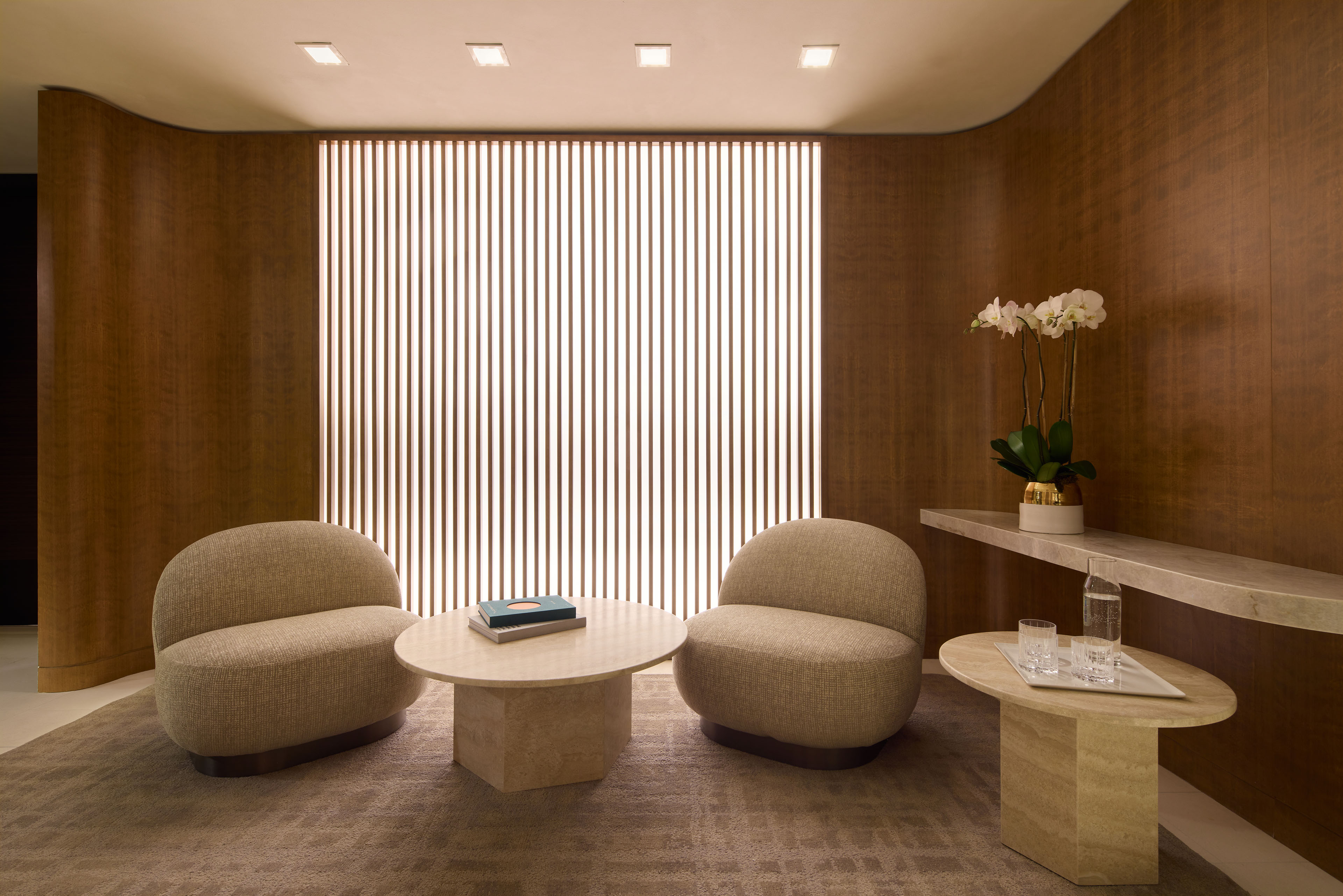 Spa lounge with curved wood-paneled walls, upholstered chairs, stone tables, orchids, and a softly illuminated slatted screen.