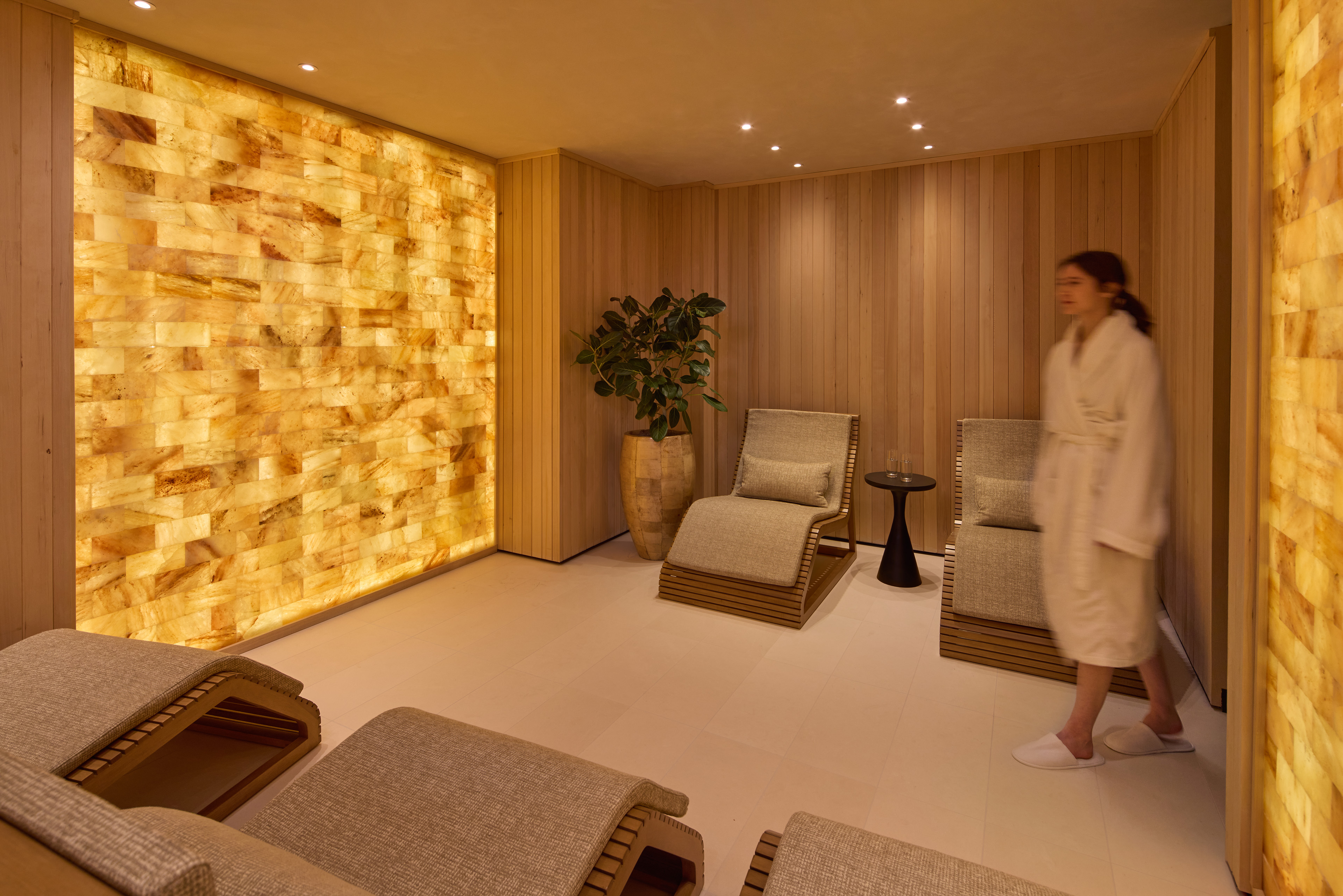 Spa relaxation room with wood-paneled walls, illuminated stone features, lounge chairs, and a guest in a robe walking through the space.