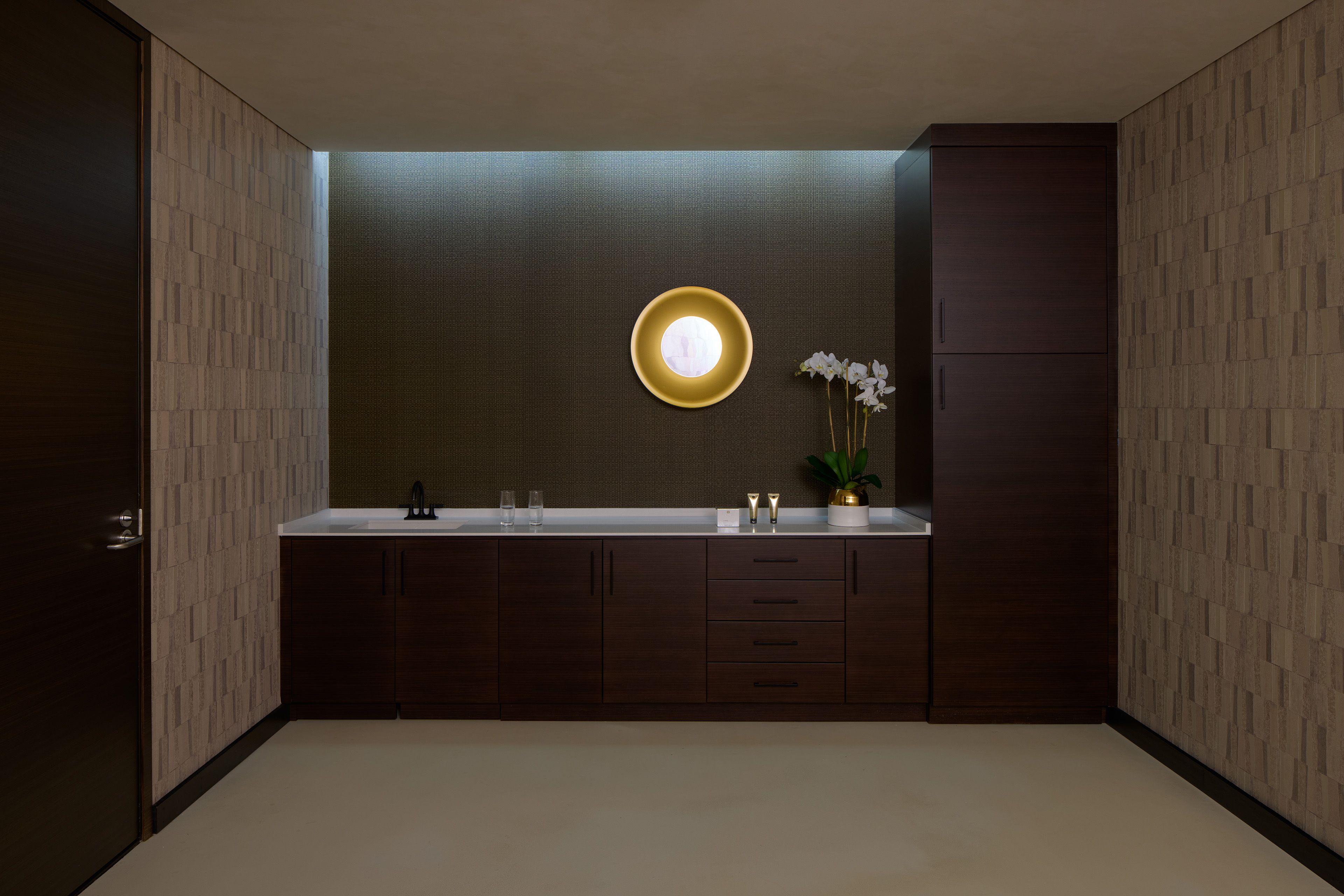 Interior of a spa treatment area with dark wood cabinetry, a long white countertop, textured wall finishes, and a circular illuminated mirror above a display of orchids.