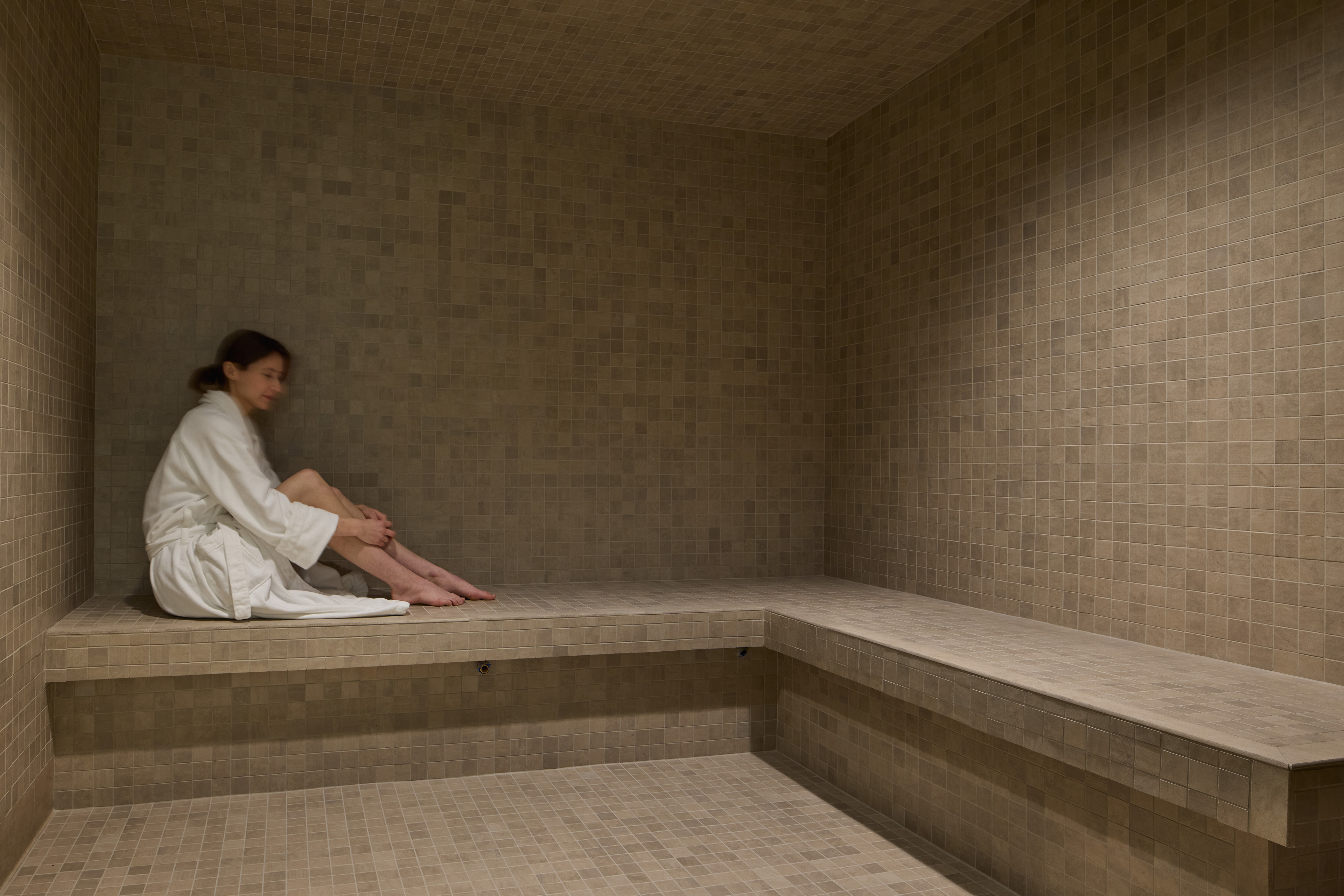 Tiled steam room with built-in benches and a guest in a white robe seated along the wall.