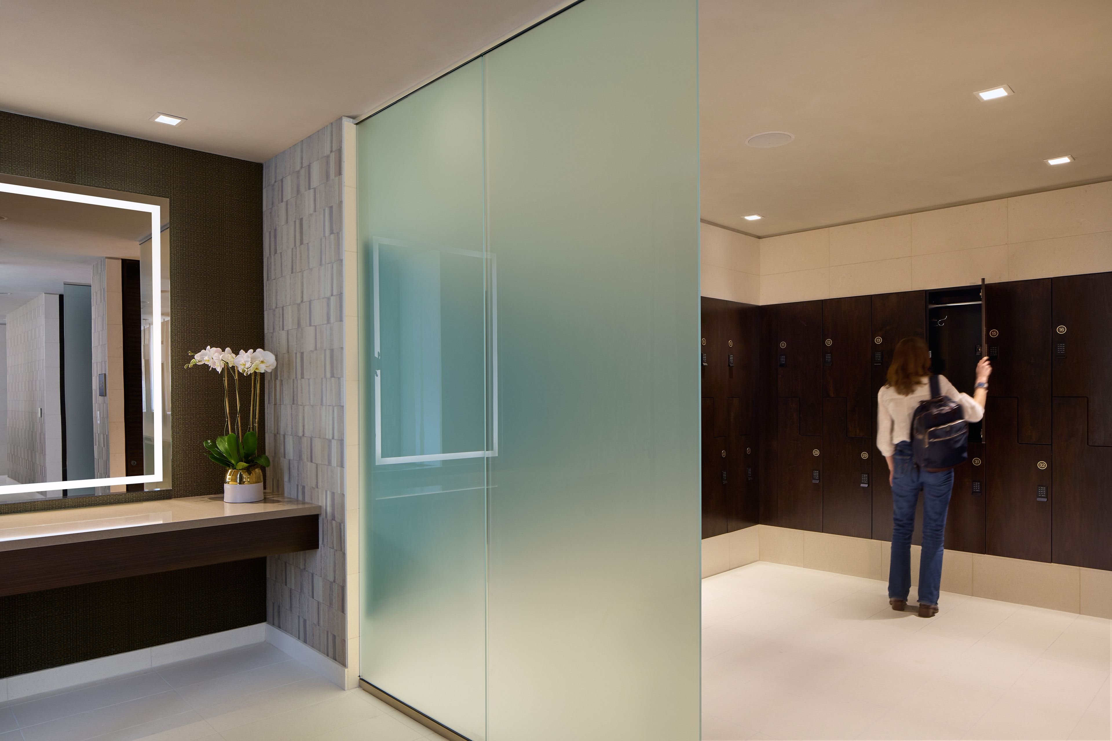 Spa locker room with dark wood lockers, an illuminated vanity mirror, orchids, and a frosted glass partition.
