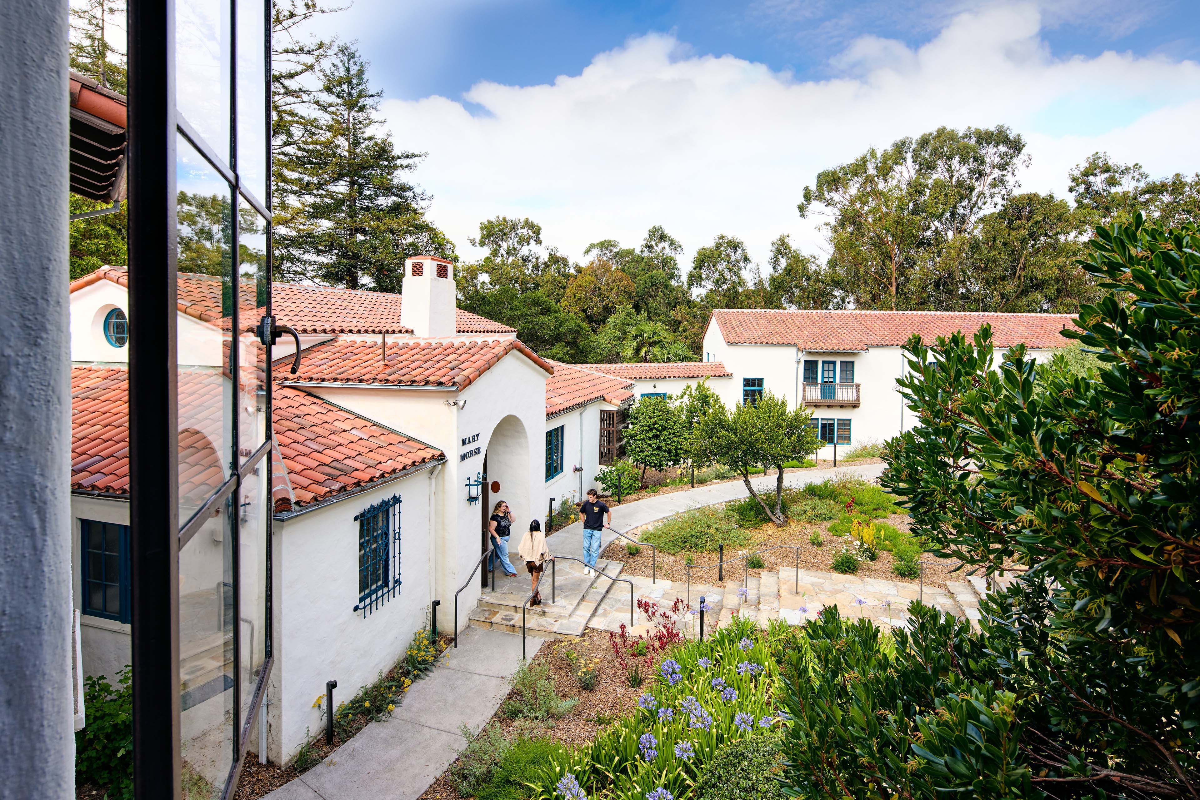 View across a landscaped courtyard of white stucco campus buildings with red tile roofs, blue-trimmed windows, mature trees, and students walking between buildings.