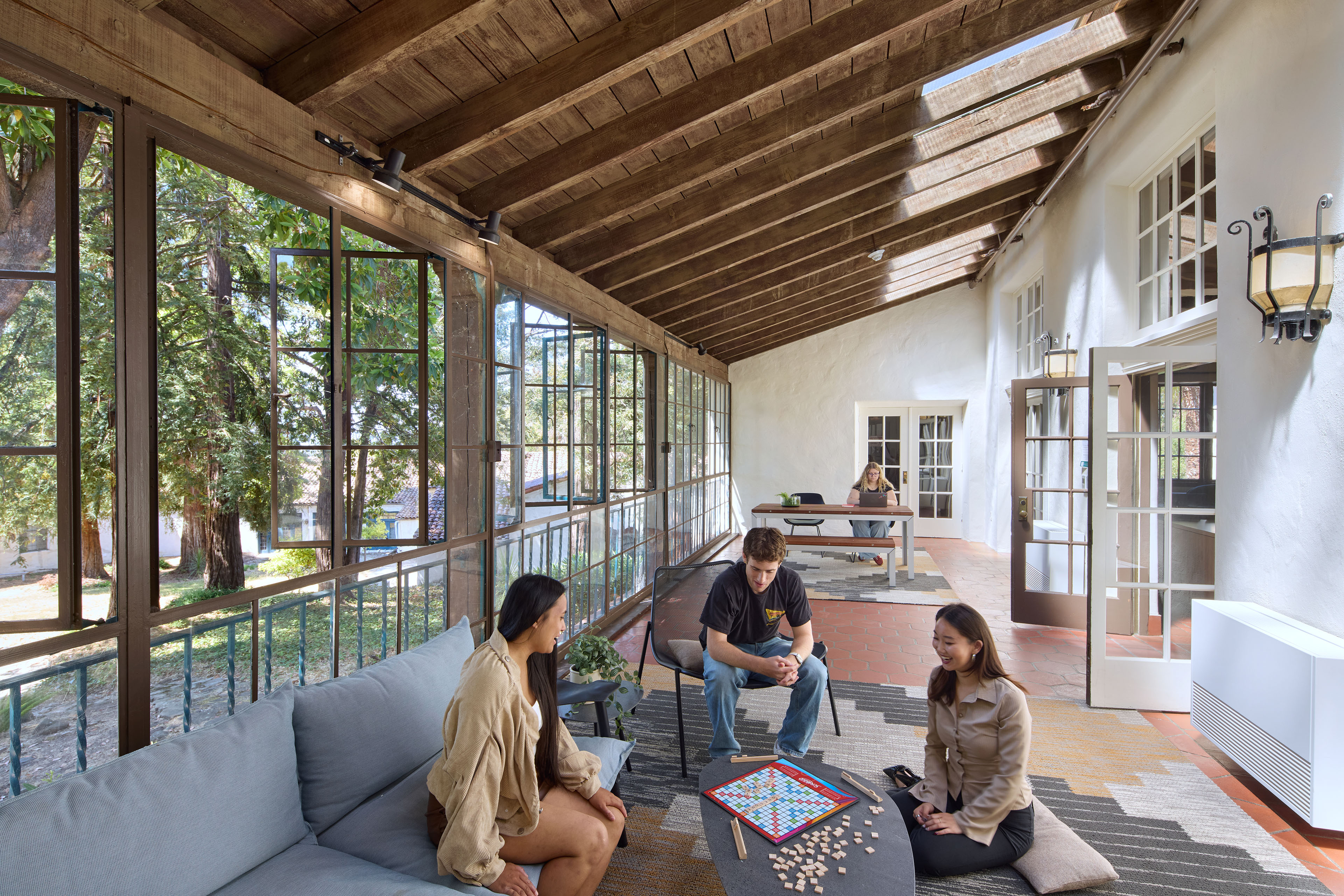 Enclosed porch with exposed wood framing, large operable windows, lounge seating, and students studying and playing a board game.