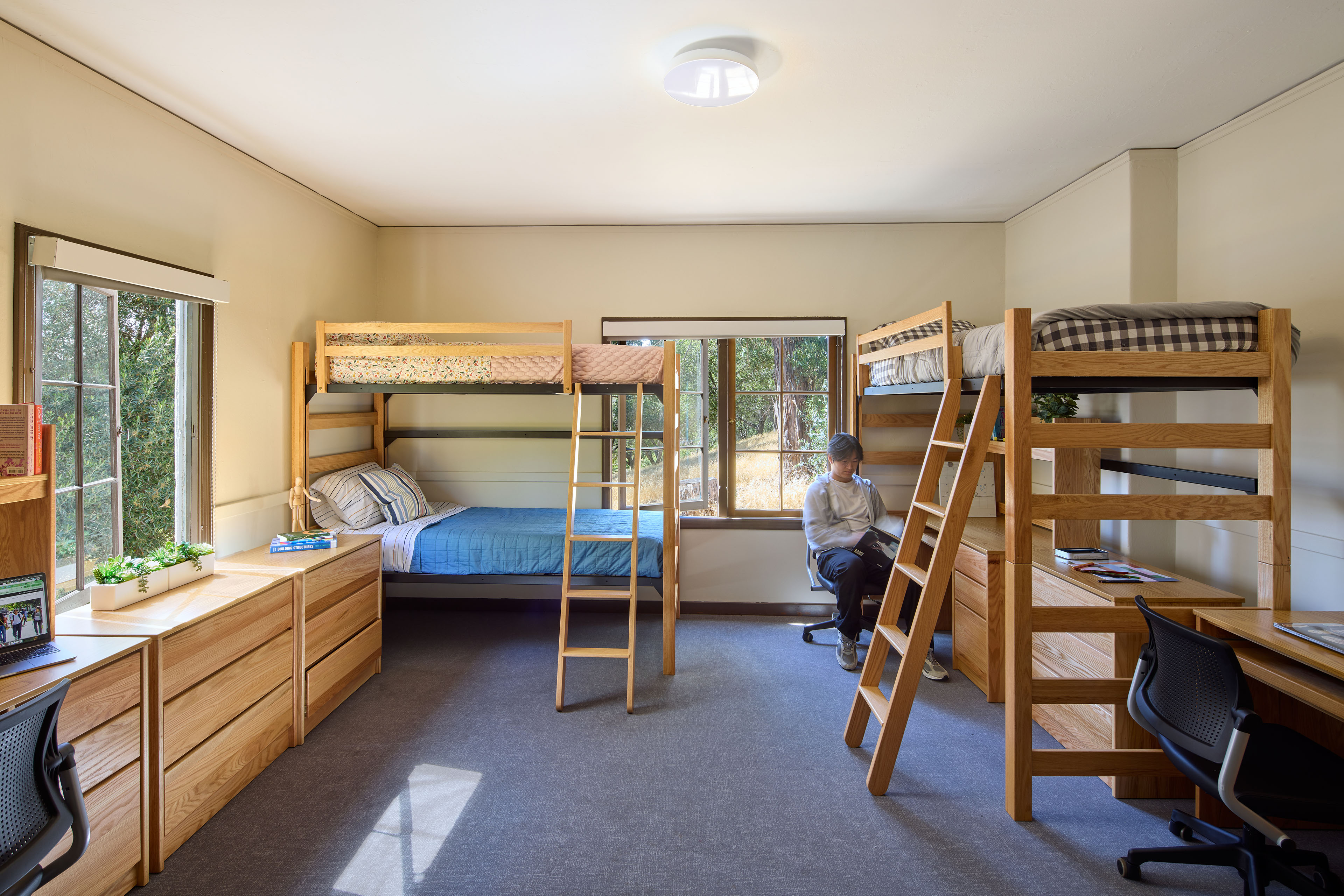 Larger residence hall room with lofted beds, desks, wood storage, and multiple windows, with a student seated at a desk.