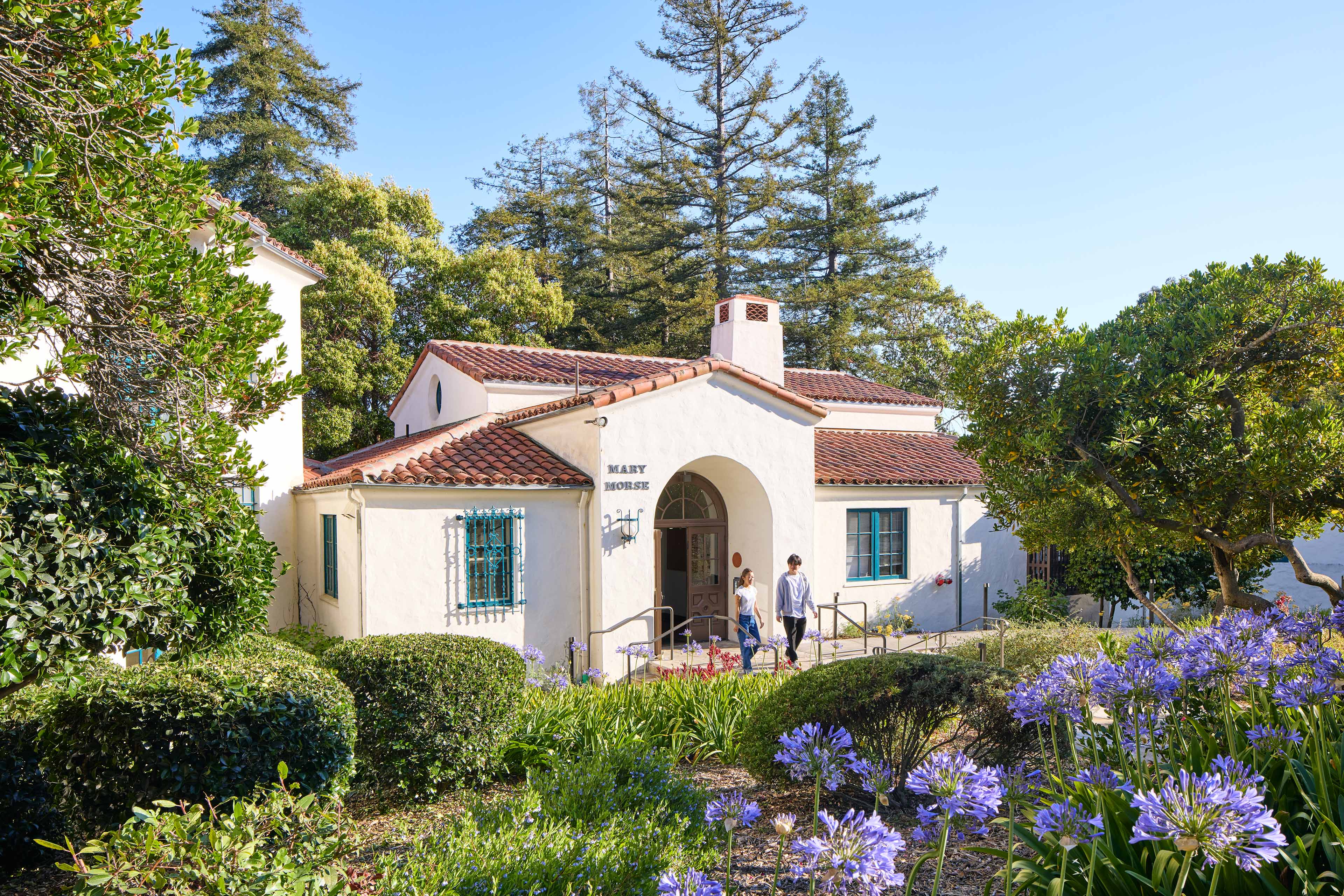 Exterior view of Mary Morse Hall with white stucco walls, red tile roofs, blue-trimmed windows, landscaped gardens, and two students walking near the arched entrance.