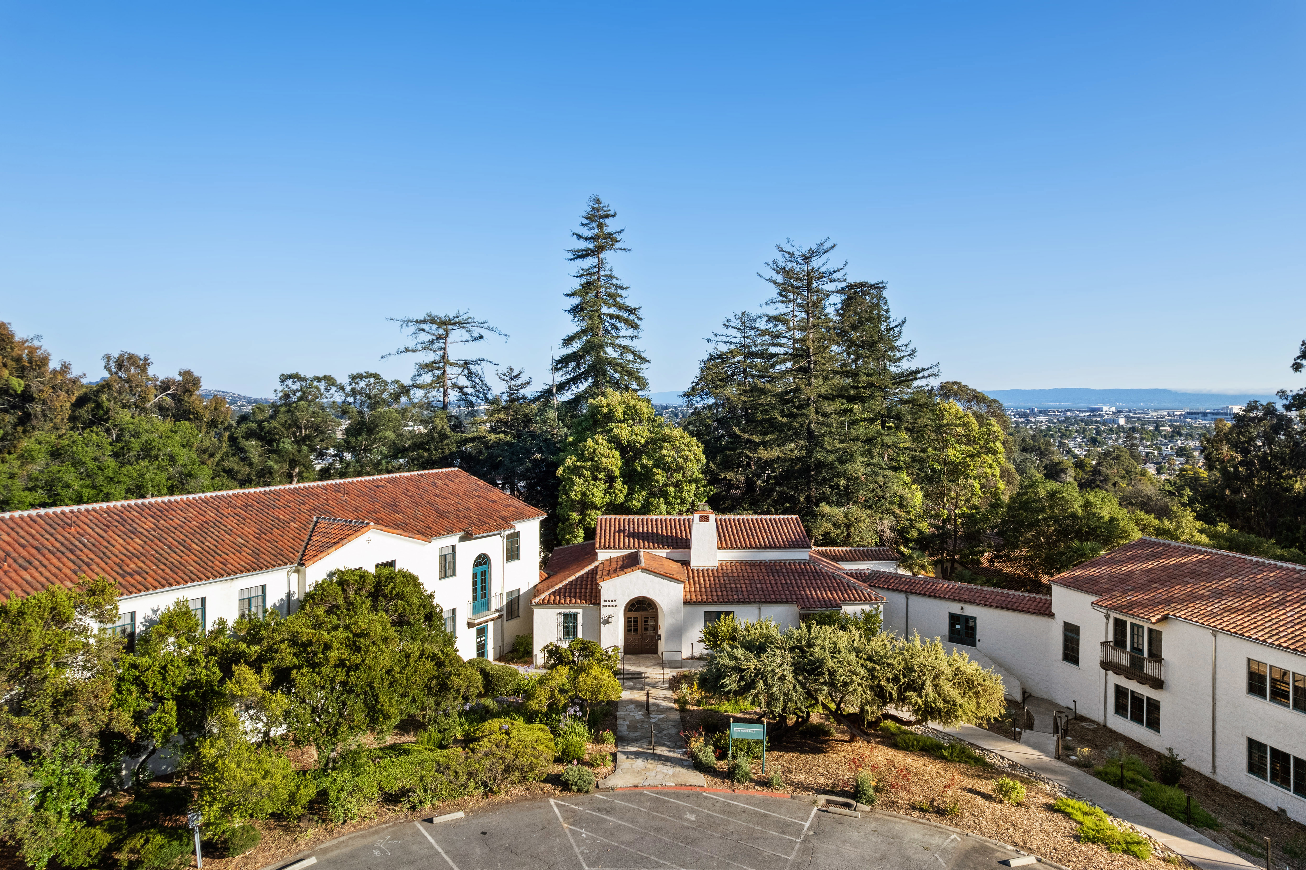 Elevated exterior view of Mary Morse Hall, showing white stucco buildings with red tile roofs surrounded by mature trees and landscaped grounds, with the city visible in the distance.