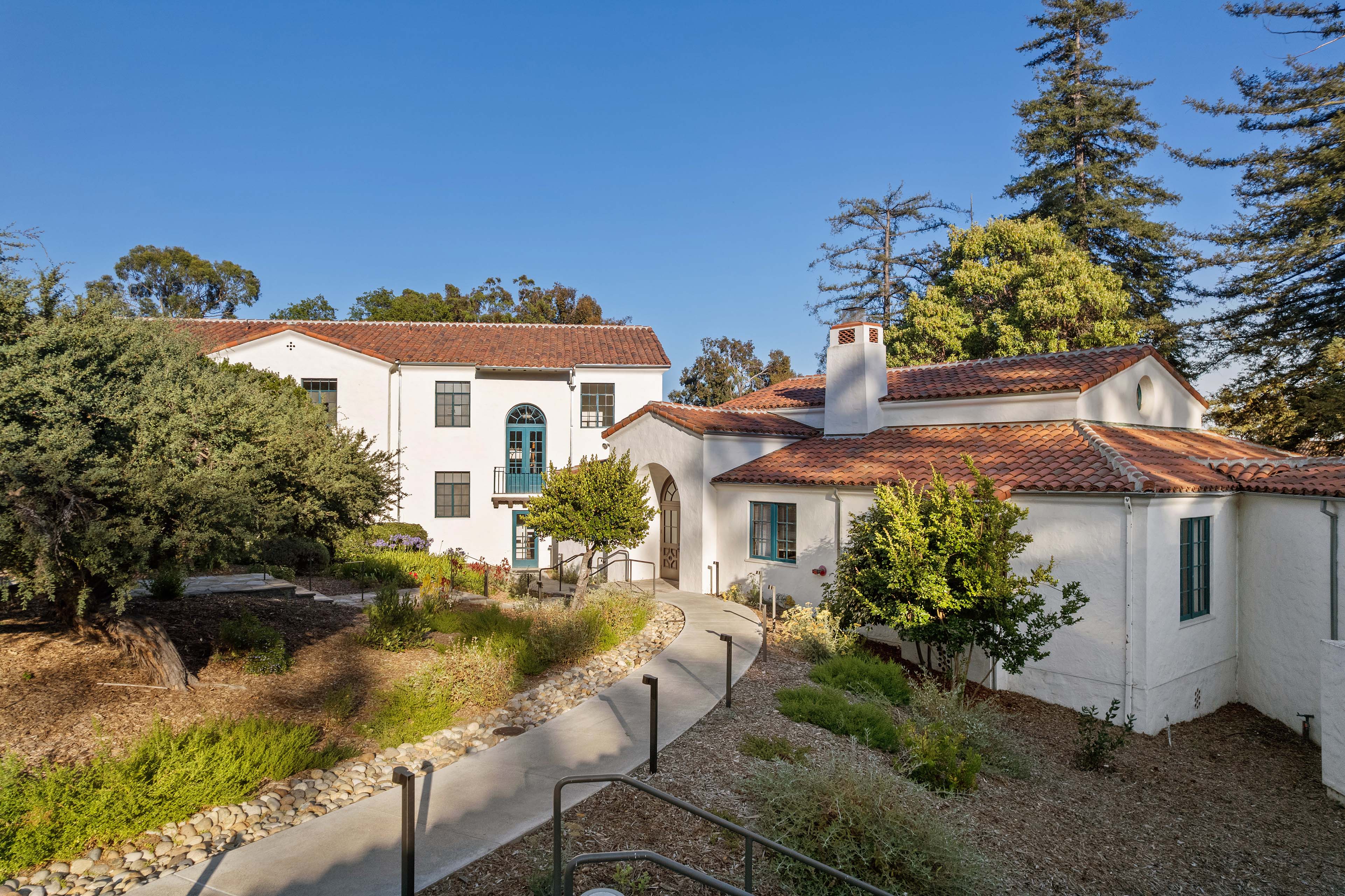 Exterior view of Mary Morse Hall with white stucco walls, red tile roofs, landscaped pathways, and mature trees under a clear blue sky.