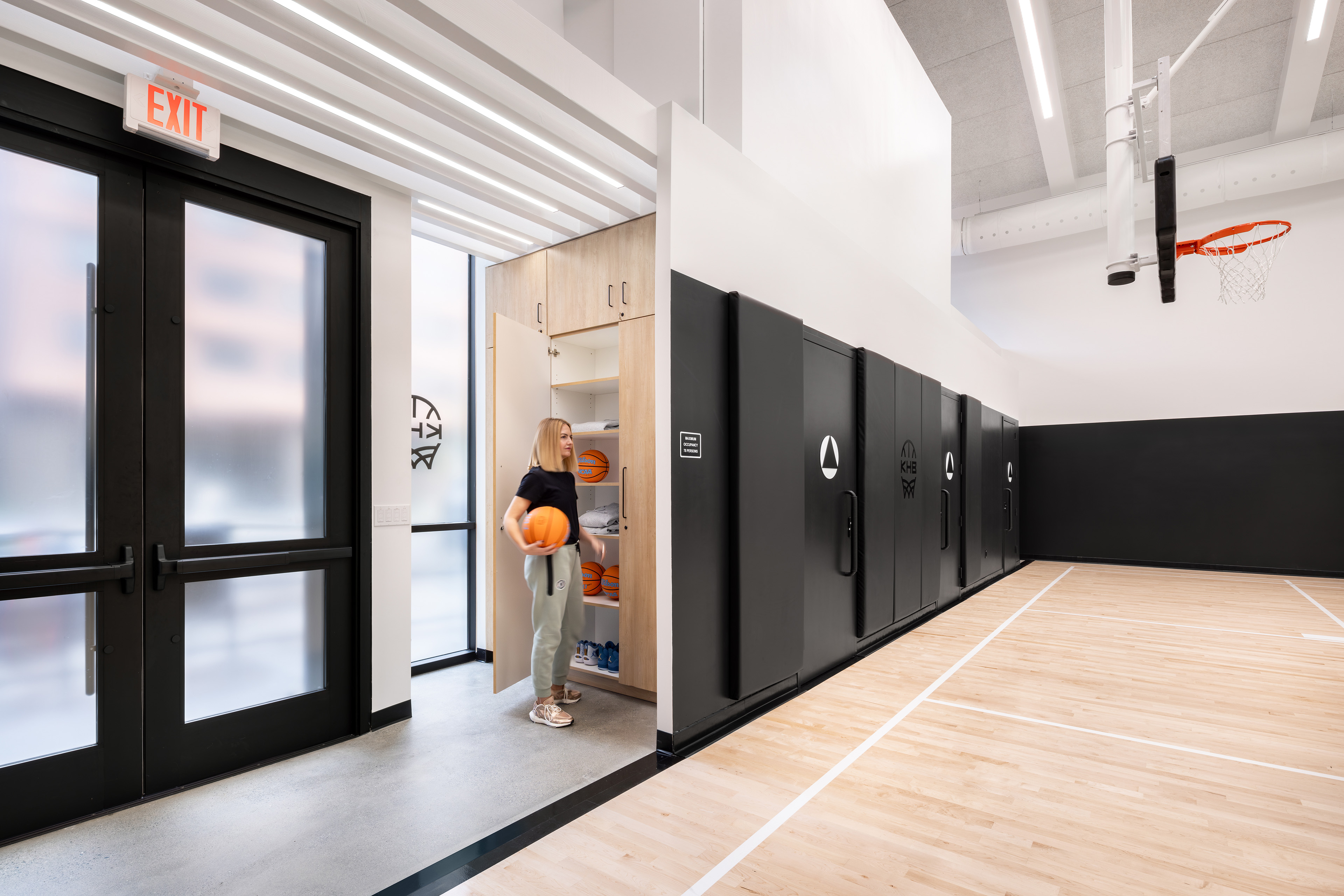 A woman stands in a modern indoor basketball facility next to a row of large, matte black storage cabinets. She is holding a basketball and opening a light-colored wooden cabinet that holds shelves filled with additional basketballs. To her right is an indoor basketball court with hardwood floors and a hoop. To her left is a set of dark-framed exit doors with frosted glass panels underneath a bright, glowing 